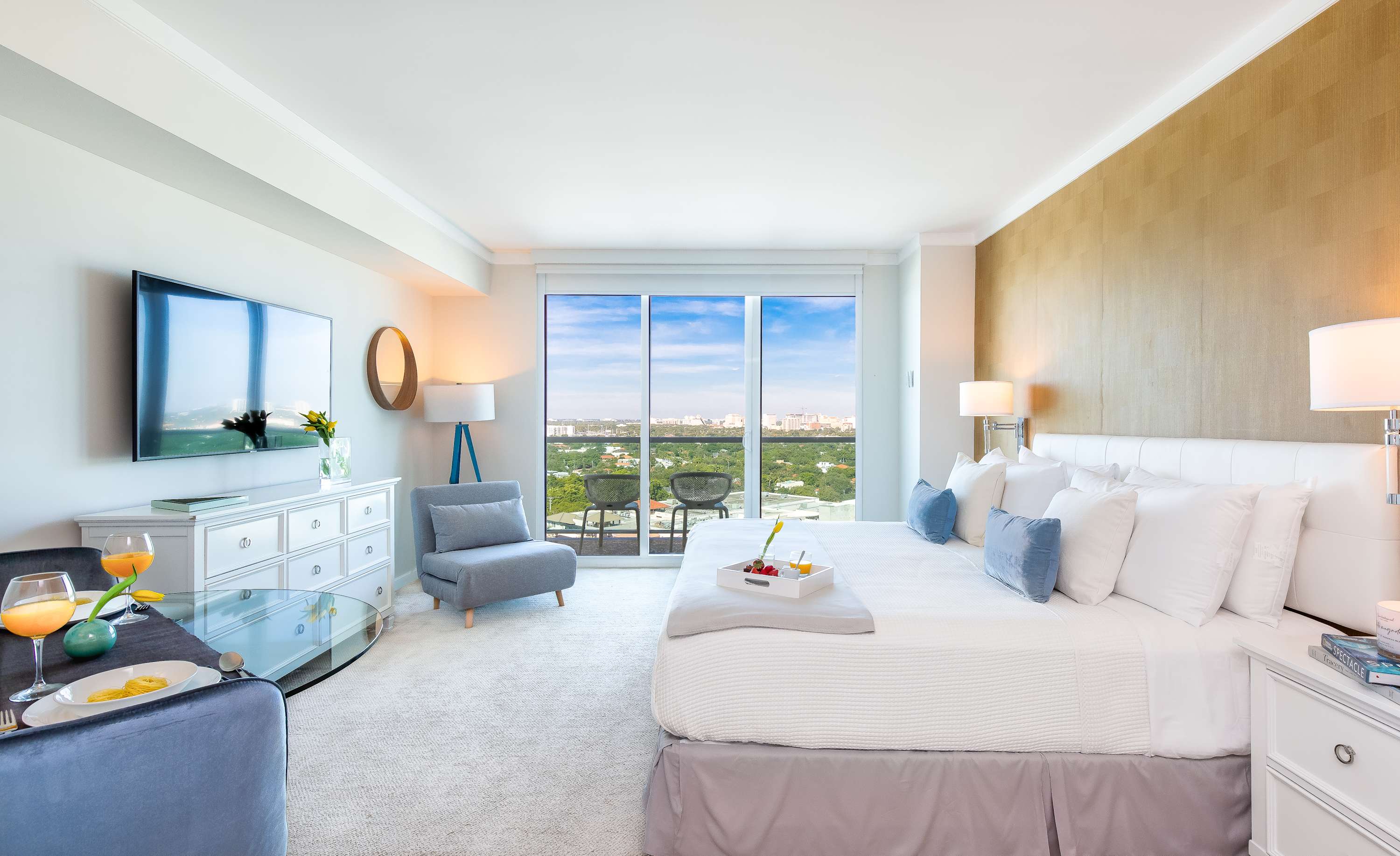 A wide shot of a bright studio apartment, highlighting a king-sized bed with white and blue linens, a glass dining table with blue chairs, and a wall-mounted flat-screen TV. A large window offers a city view, and the room is carpeted with light-colored walls.