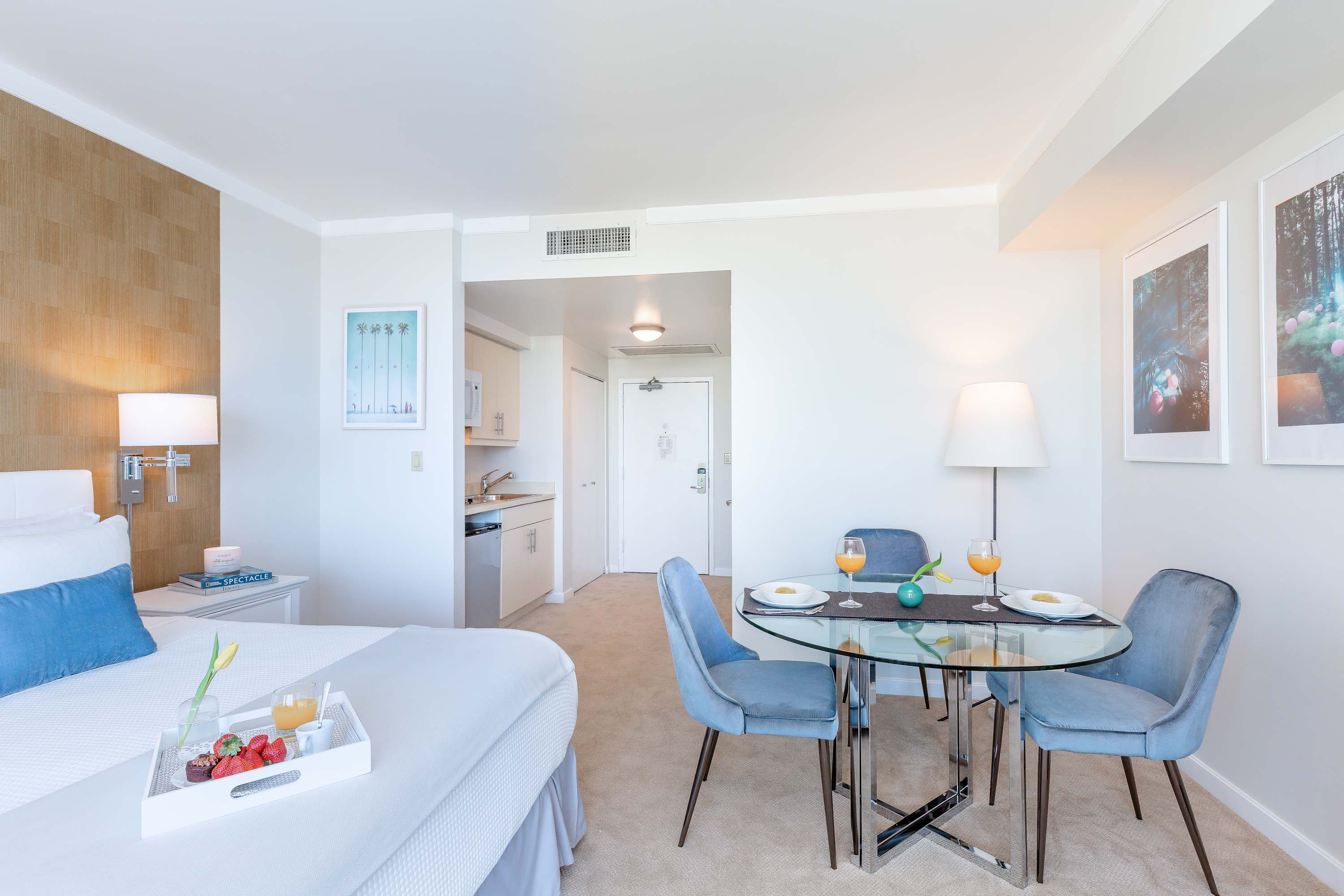 A bright studio apartment showcasing a king-sized bed with white and blue bedding, a glass-top dining table with blue velvet chairs, and a compact kitchenette in the background. The room features light walls, carpeted flooring, and a textured accent wall.