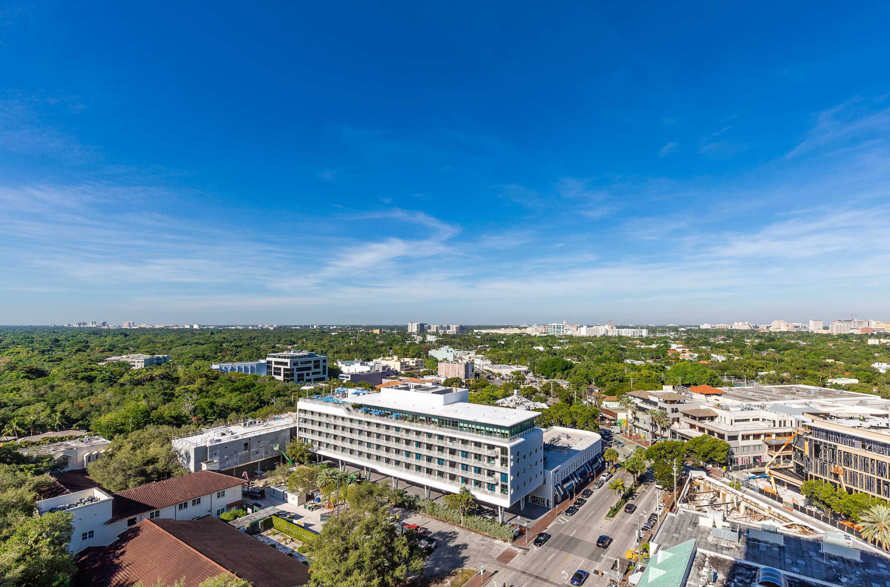 An expansive aerial view of a city with numerous buildings and lush green trees under a clear blue sky with scattered white clouds. A multi-story building with a rooftop pool is visible in the mid-ground, surrounded by other urban structures and dense foliage.