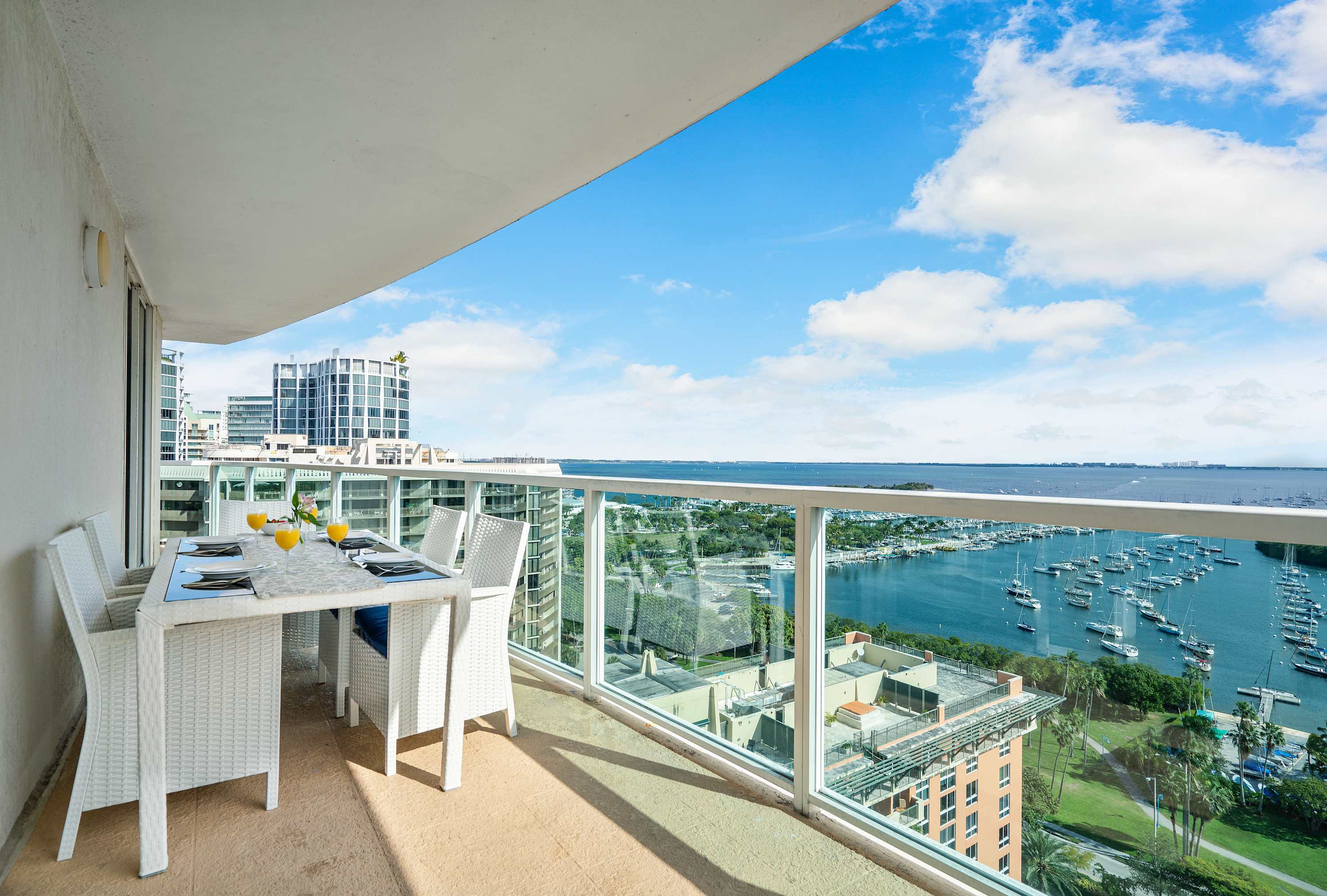 Spacious outdoor balcony with a white wicker dining set for four, offering panoramic views of the bay, marina with boats, and distant city skyline under a bright blue sky.