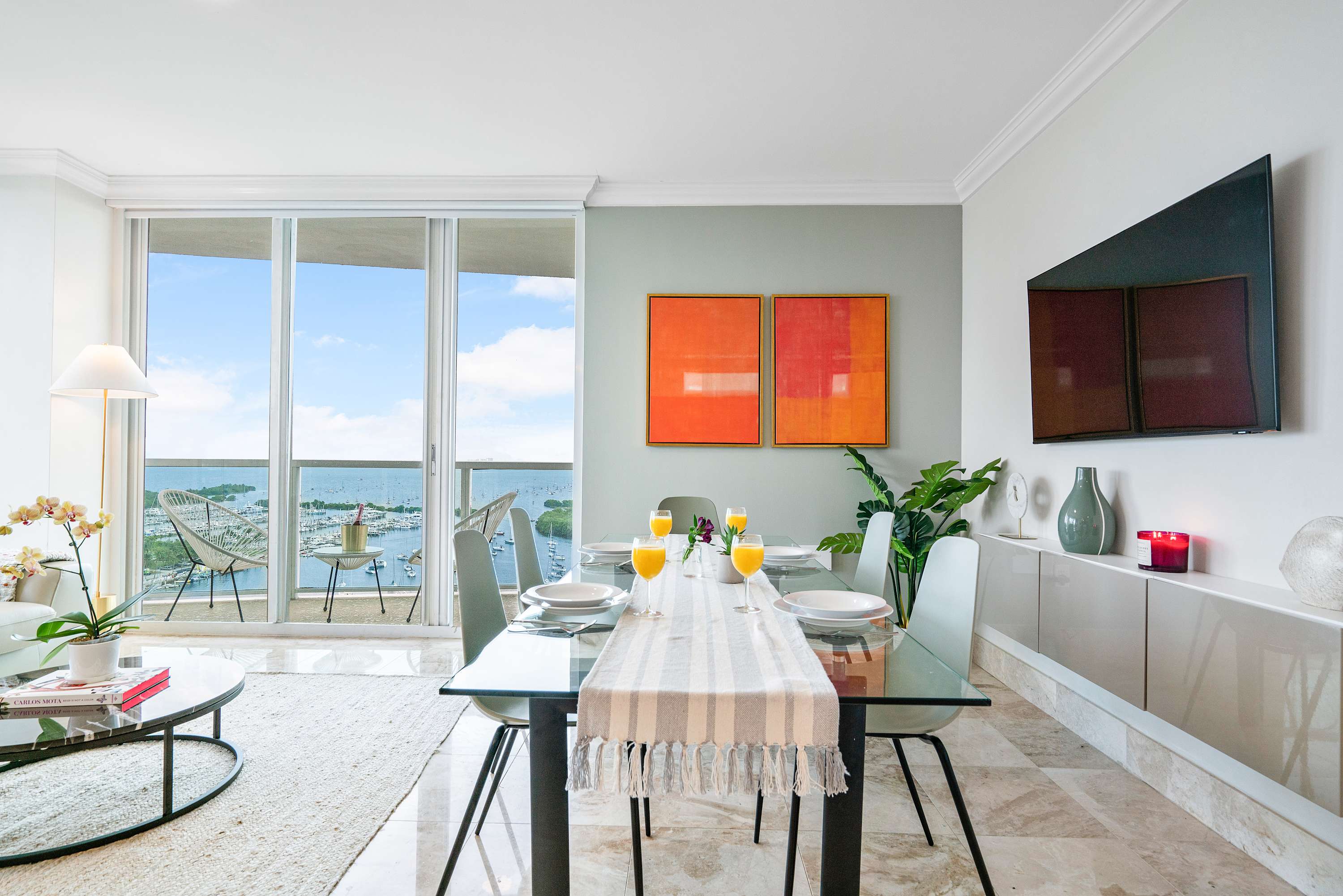Modern dining area with a glass-top table and light gray chairs, adjacent to a living room with a white sofa, all set against large windows offering expansive bay and city views.