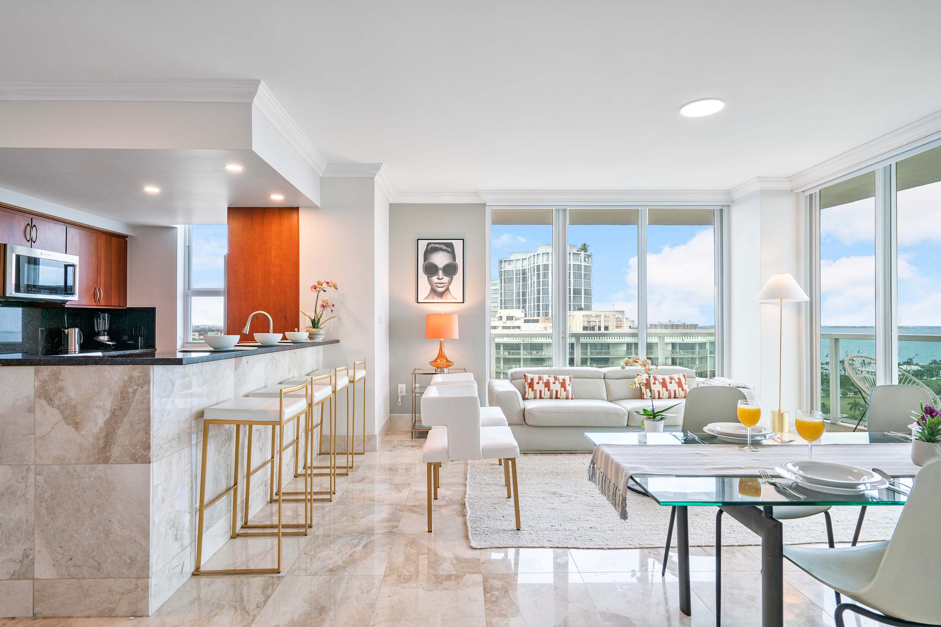 Open-concept living space with light-colored furniture, marble-patterned tile floors, and a dining area, featuring a kitchen bar with stools and large windows offering city and bay views.