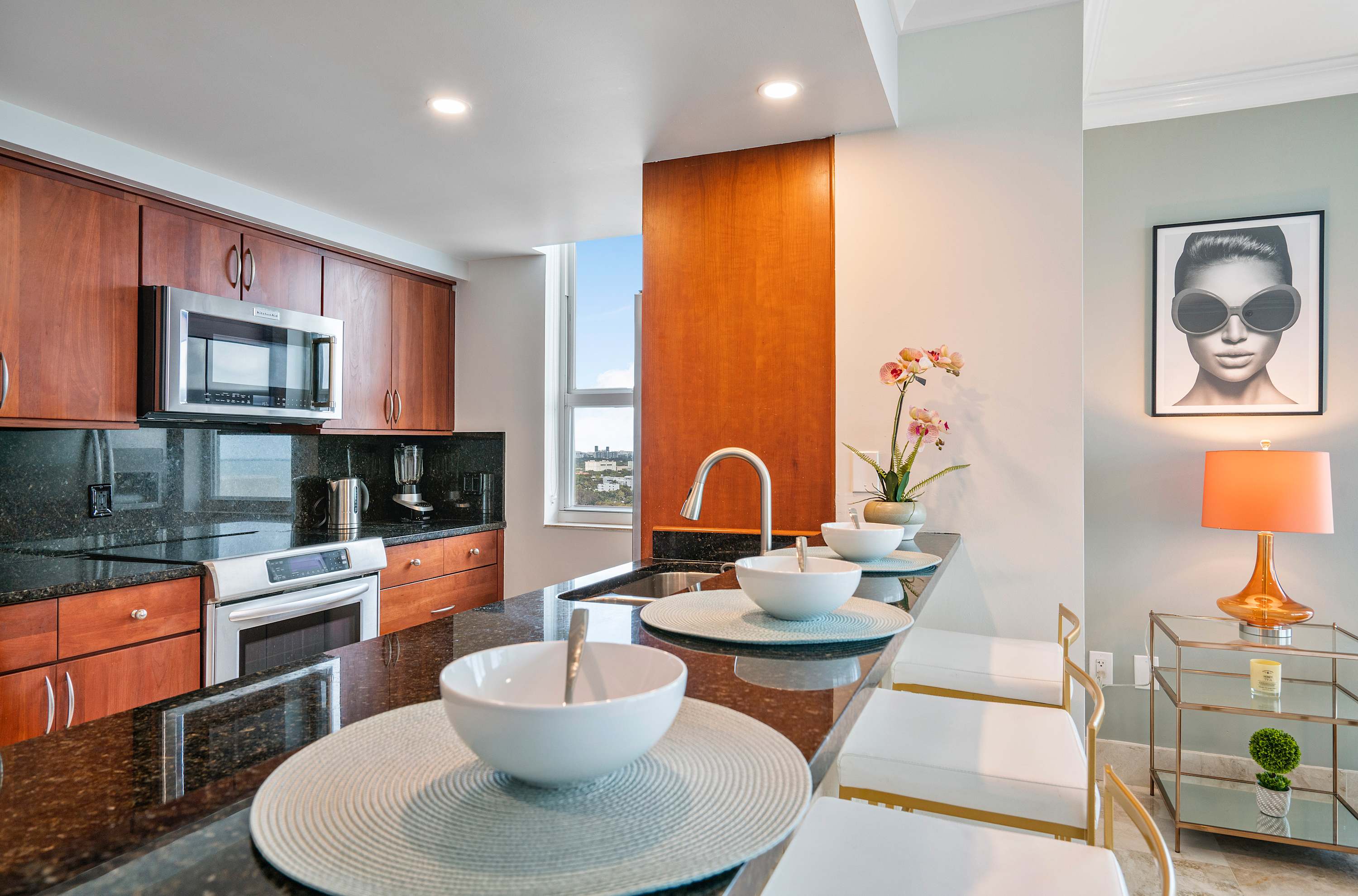 Modern kitchen with dark granite countertops, rich wooden cabinetry, and stainless steel appliances, featuring a breakfast bar with three white and gold stools.