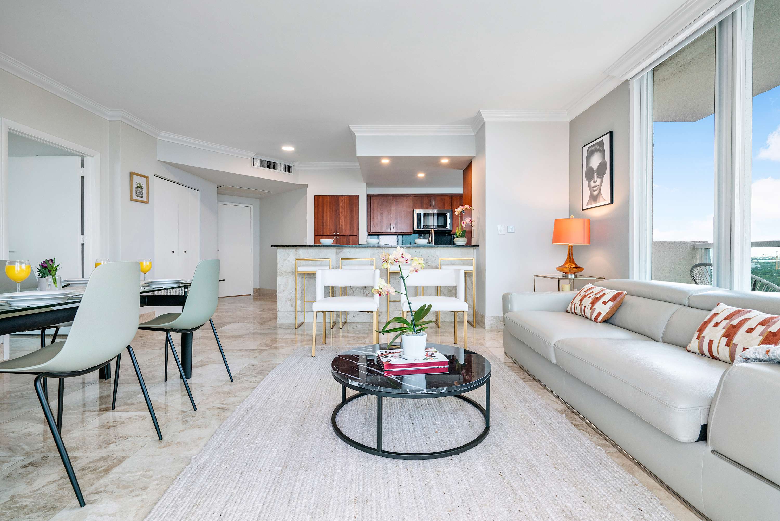 Open-concept living room with a white leather sofa and armchairs, facing a dining area and a kitchen bar, all featuring marble-patterned tile floors and bright natural light.
