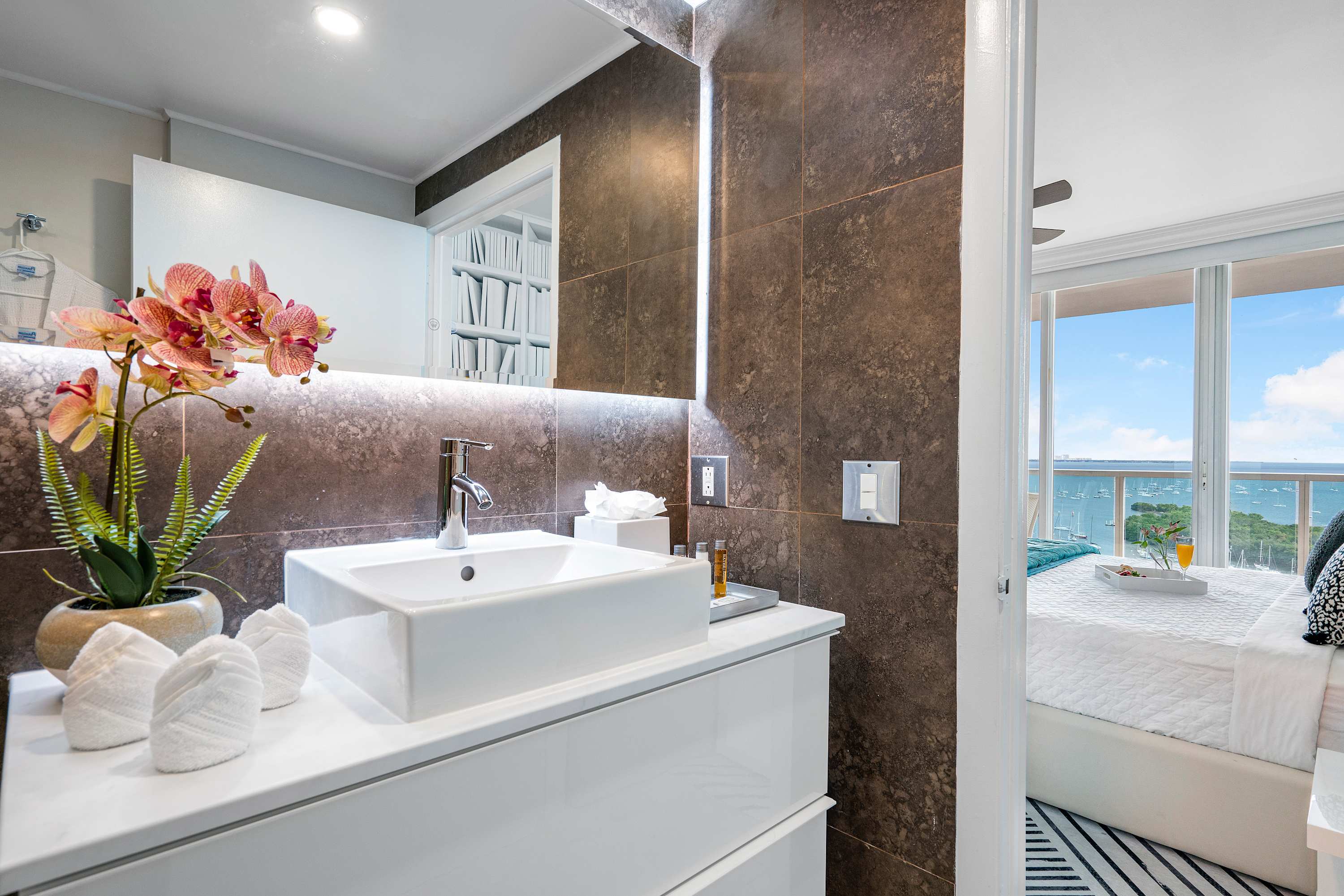 A close-up of the modern bathroom vanity with a white rectangular sink, chrome faucet, and a large mirror with vertical LED lighting. Through the open doorway, a glimpse of the bedroom with a bed and ocean view is visible.
