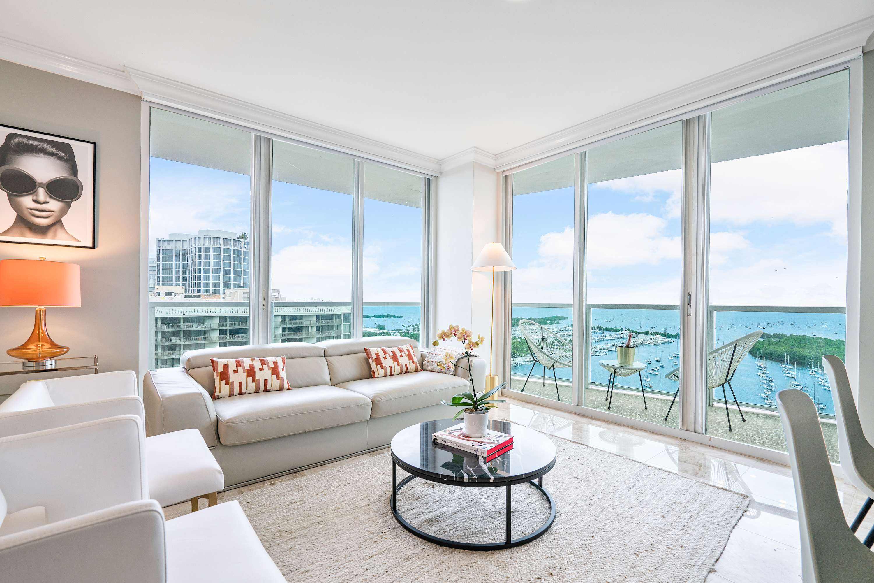 Bright living room with a white leather sofa and two white armchairs, centered around a black marble coffee table, with floor-to-ceiling windows providing stunning bay and city views.