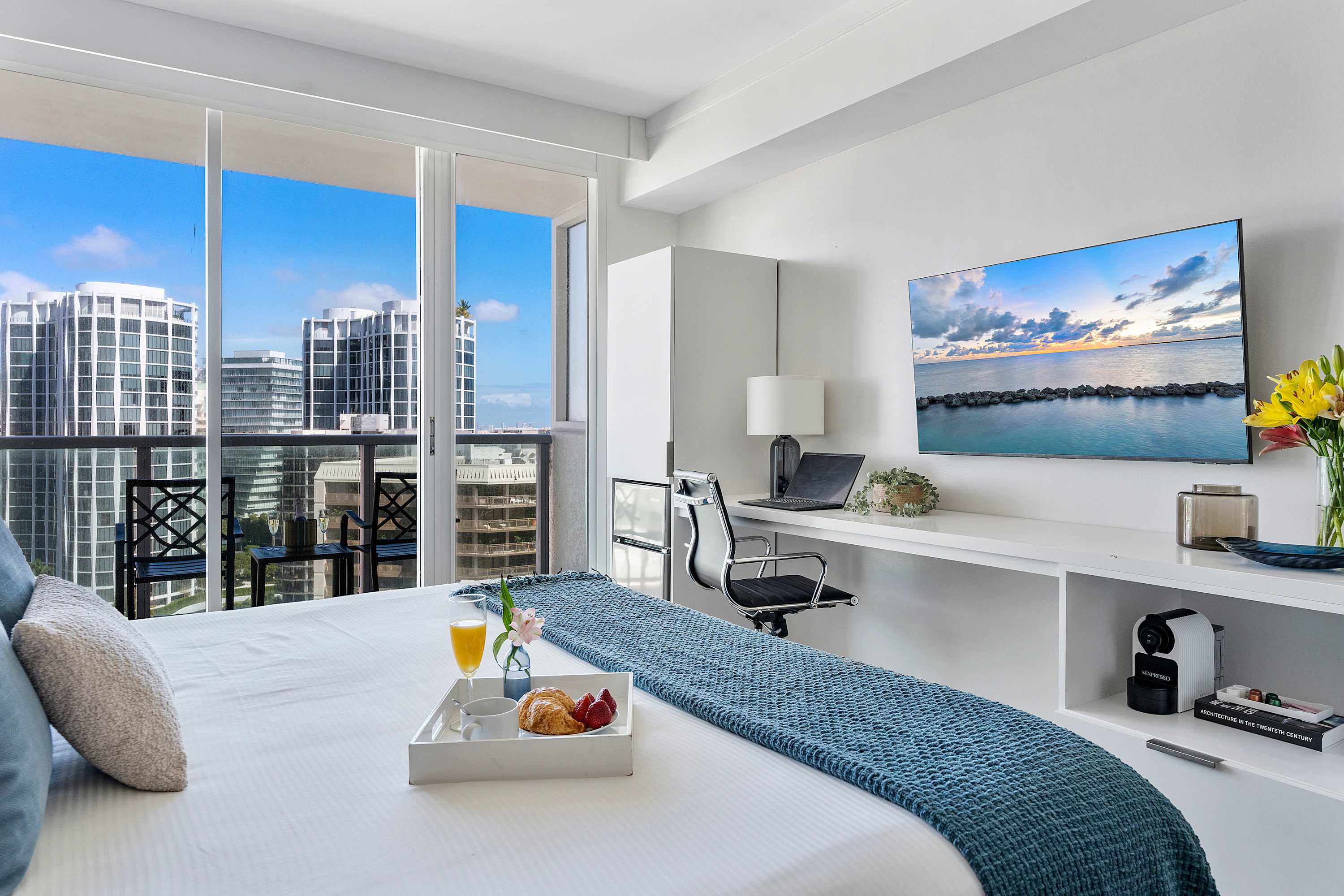 A bright studio room featuring a queen bed with a blue throw, a long white desk with a TV and coffee machine, and floor-to-ceiling windows offering city and bay views. A breakfast tray rests on the bed.