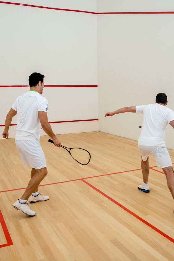 Two men in white athletic wear play squash on a brightly lit indoor court with light wooden floors and white walls marked with red lines.