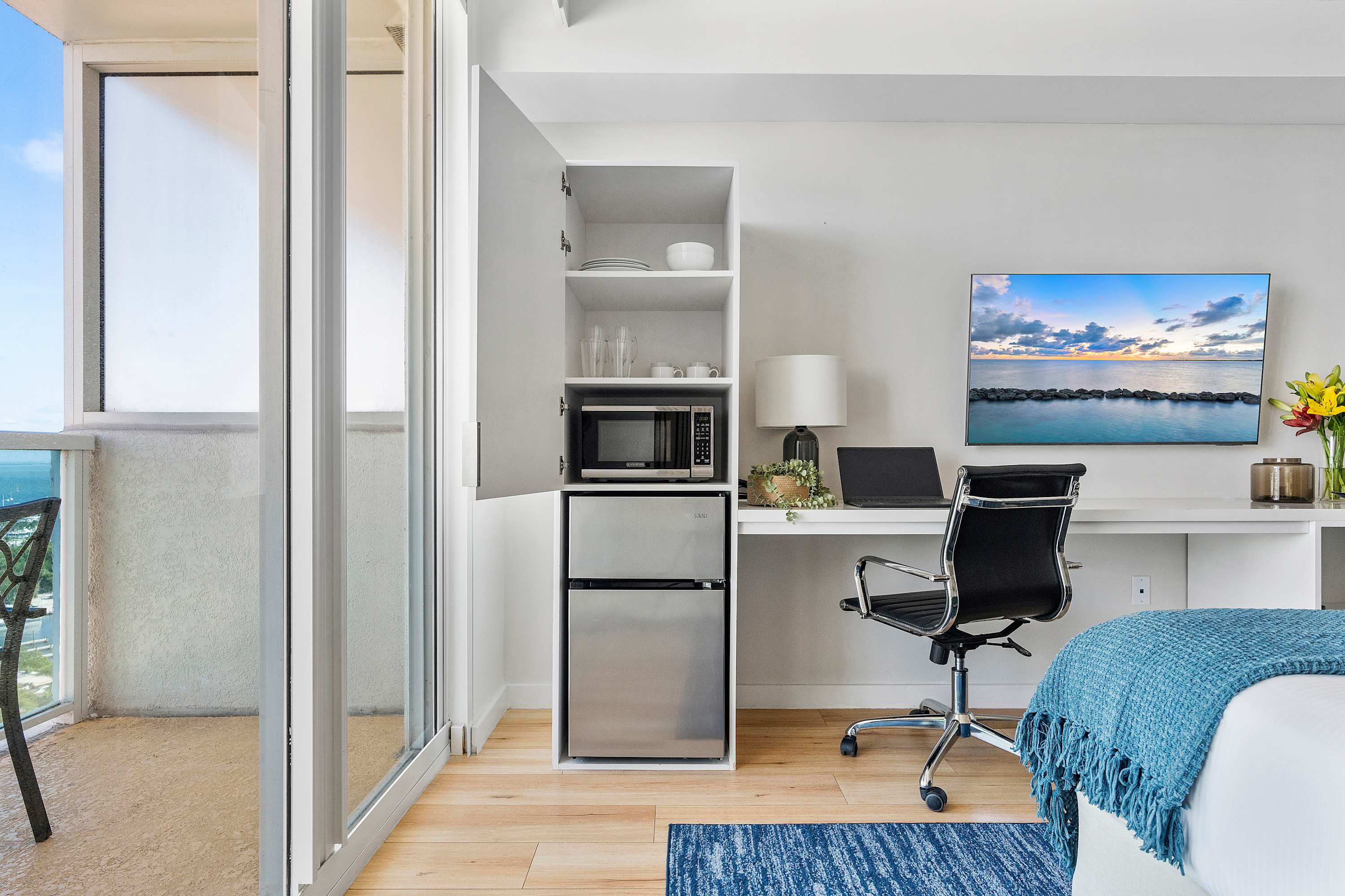 A compact kitchenette area featuring a microwave, mini-fridge, and open shelving with dishes, integrated into a white cabinet next to a modern desk with a laptop and office chair. A glass door leads to a balcony.