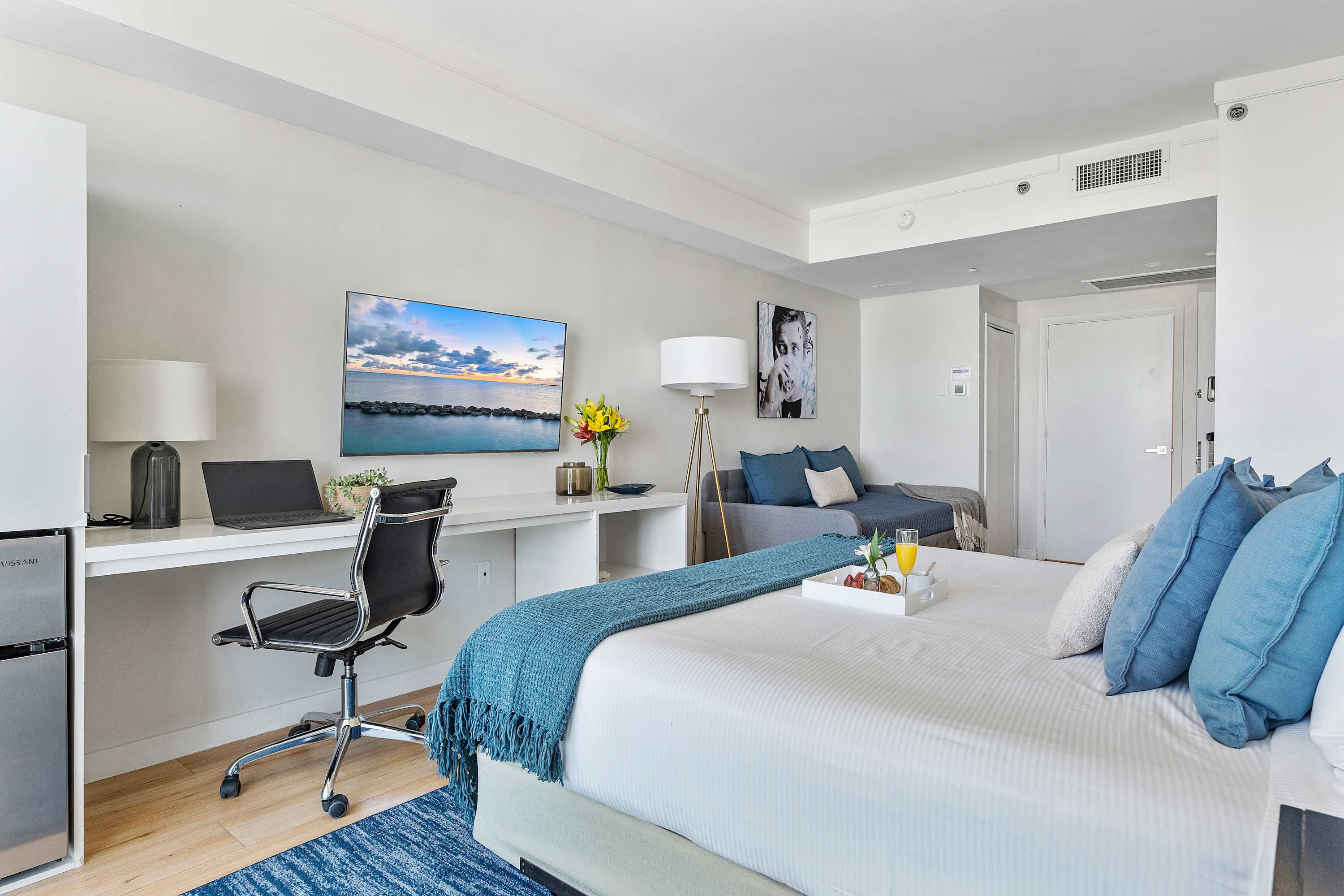 A modern studio space with a queen bed, a long white desk equipped with a laptop and TV, and a mini-fridge. The room features light wood flooring and a large abstract art piece on the wall.