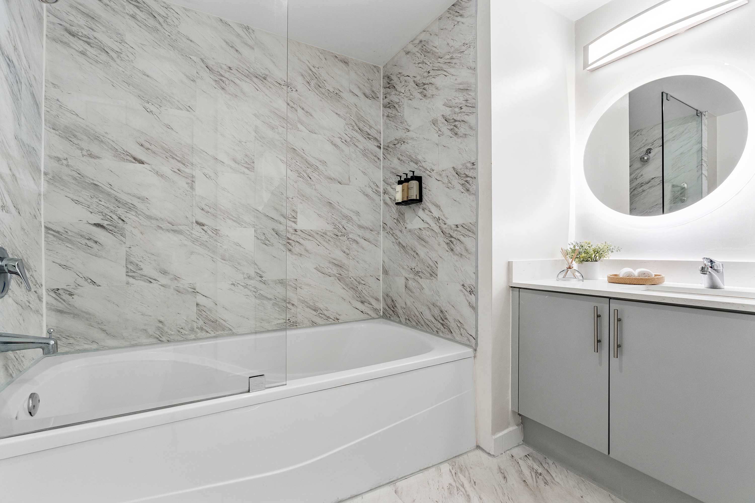 Modern bathroom featuring a white bathtub with a glass shower screen, marble-patterned wall tiles, and a vanity with a circular backlit mirror and gray cabinets.