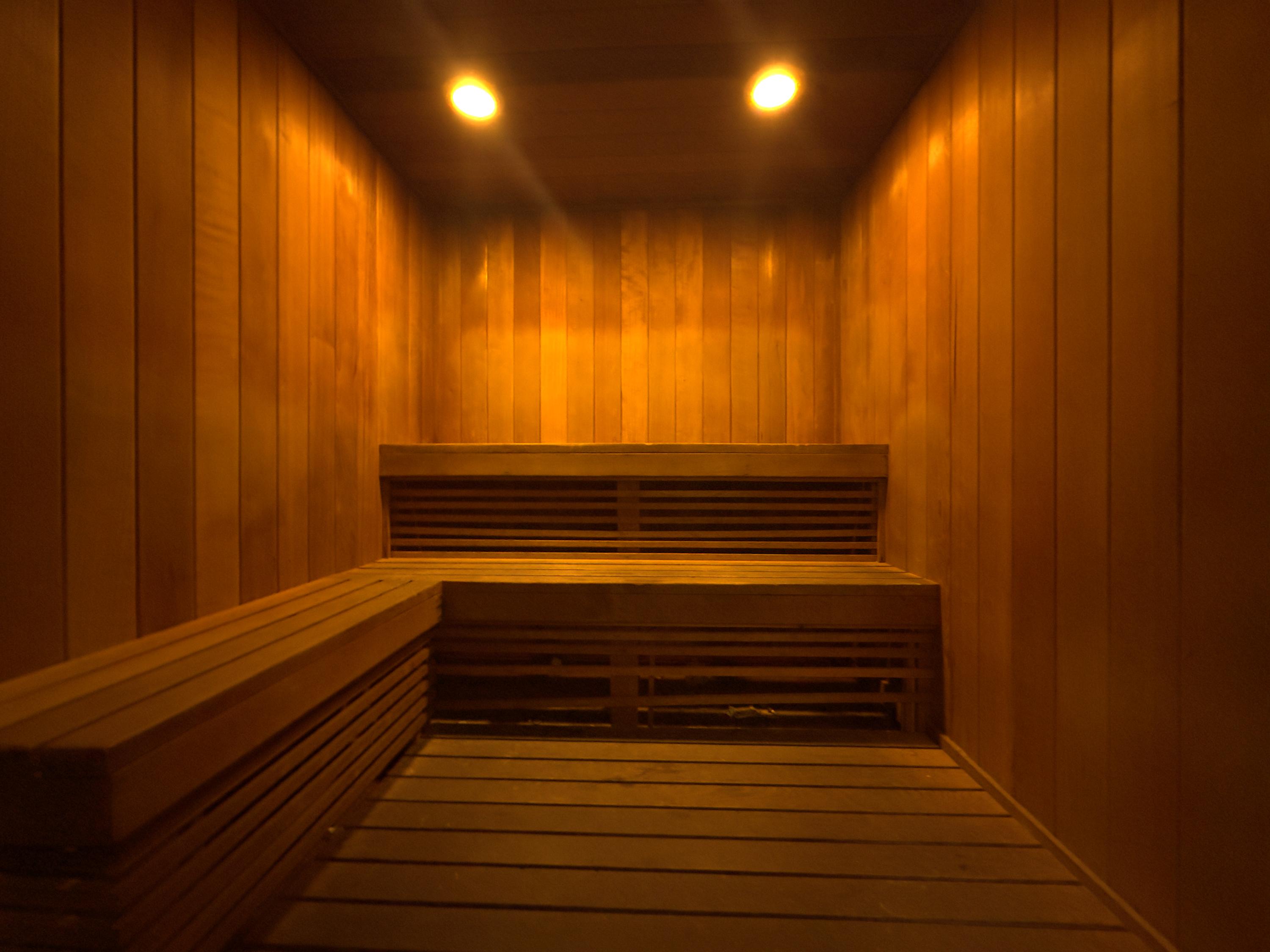 A warm, inviting sauna room features light wooden slatted walls and benches, illuminated by two bright overhead lights, creating a cozy and relaxing atmosphere.