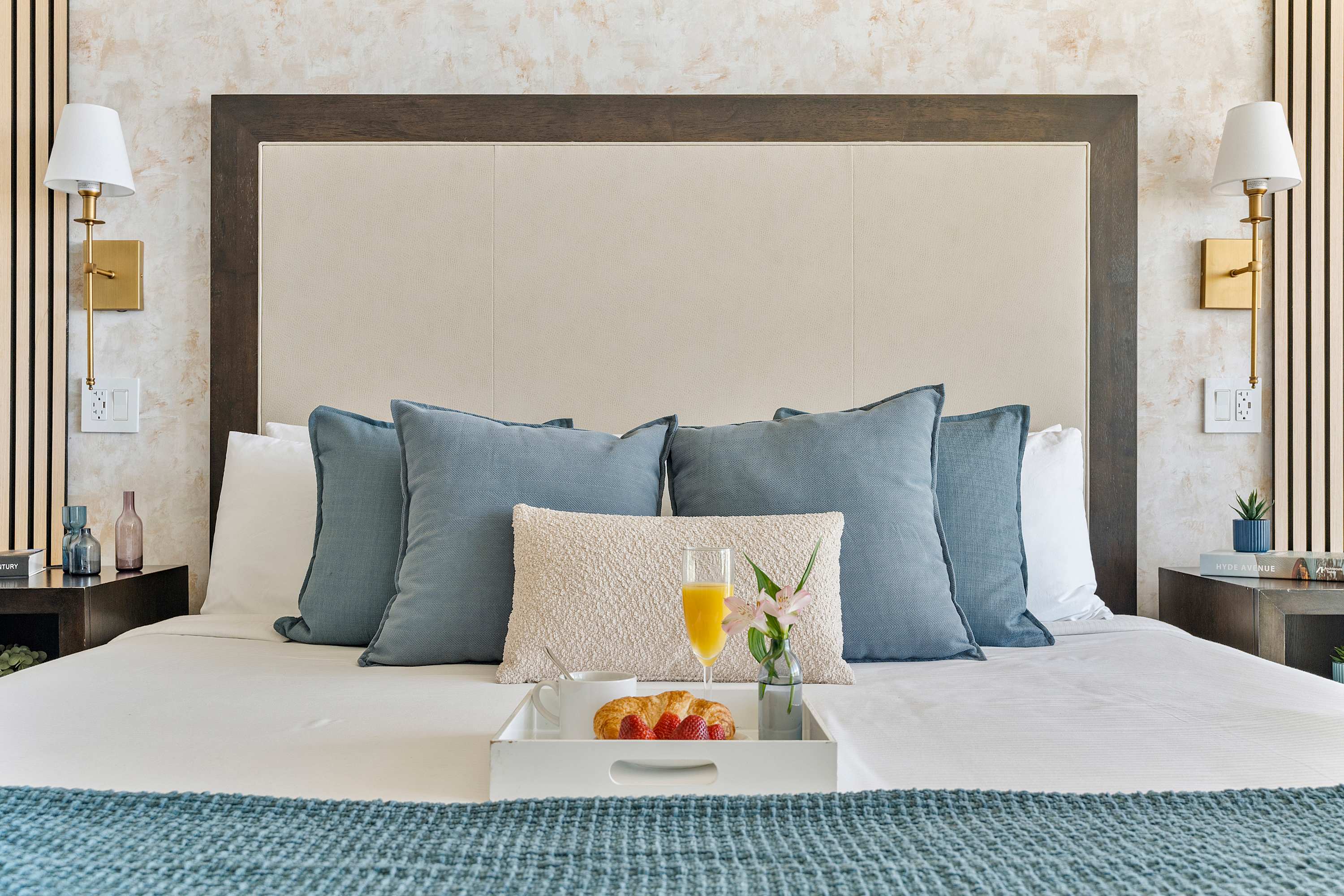 A close-up of a neatly made queen bed with a large upholstered headboard, adorned with multiple blue and cream pillows, and a breakfast tray holding orange juice, a croissant, and strawberries. Two modern wall sconces flank the bed.