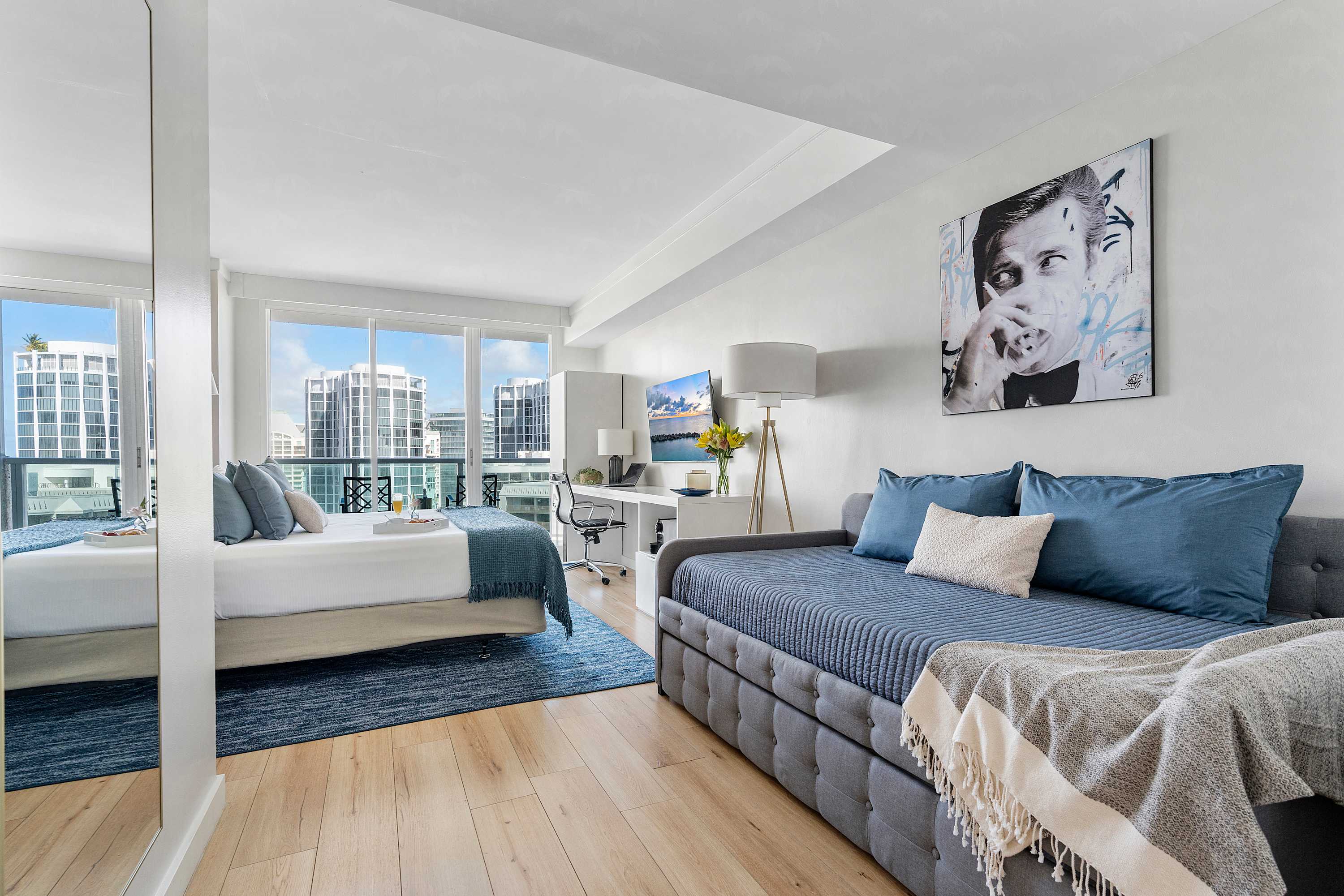 A spacious studio apartment featuring a queen bed and a grey daybed with blue pillows, a long white desk, and large windows offering city views. The room has light wood flooring and a large abstract portrait on the wall.