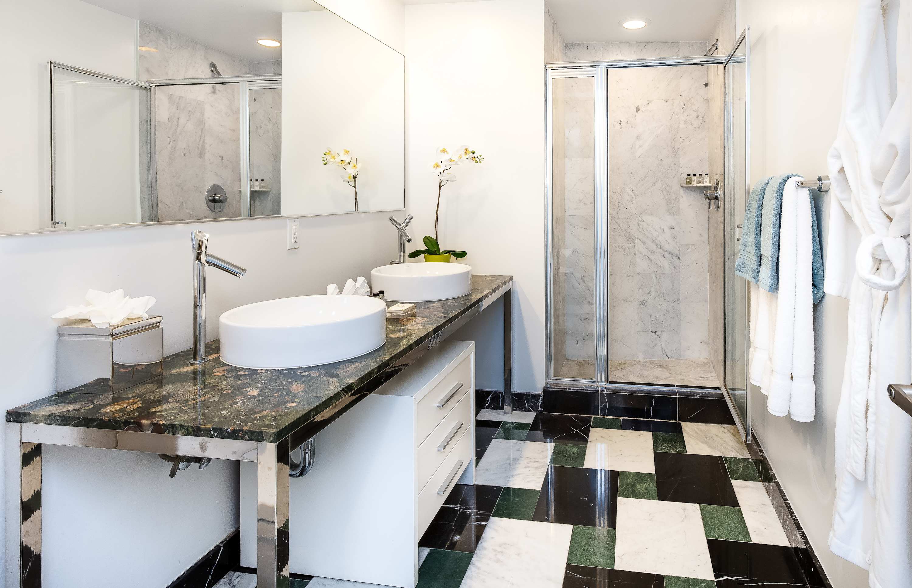 Contemporary bathroom featuring a double vanity with round white sinks set on a dark green marble countertop, a large mirror, and a walk-in glass shower. The floor has a distinctive black, white, and green checkered marble pattern.