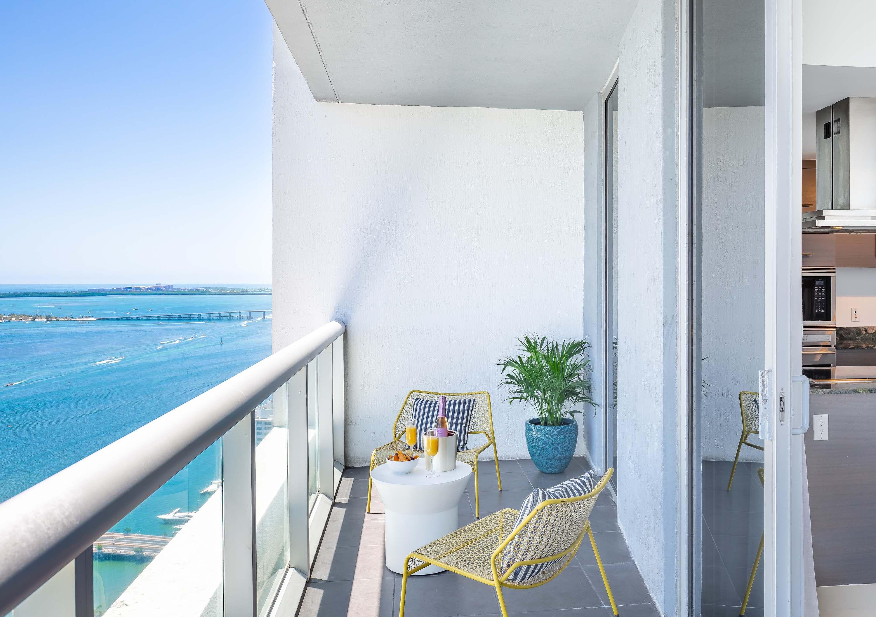 Sunny balcony with two yellow woven chairs and a white round table, offering panoramic views of the blue ocean and a distant bridge under a clear sky. A potted plant adds a touch of greenery.