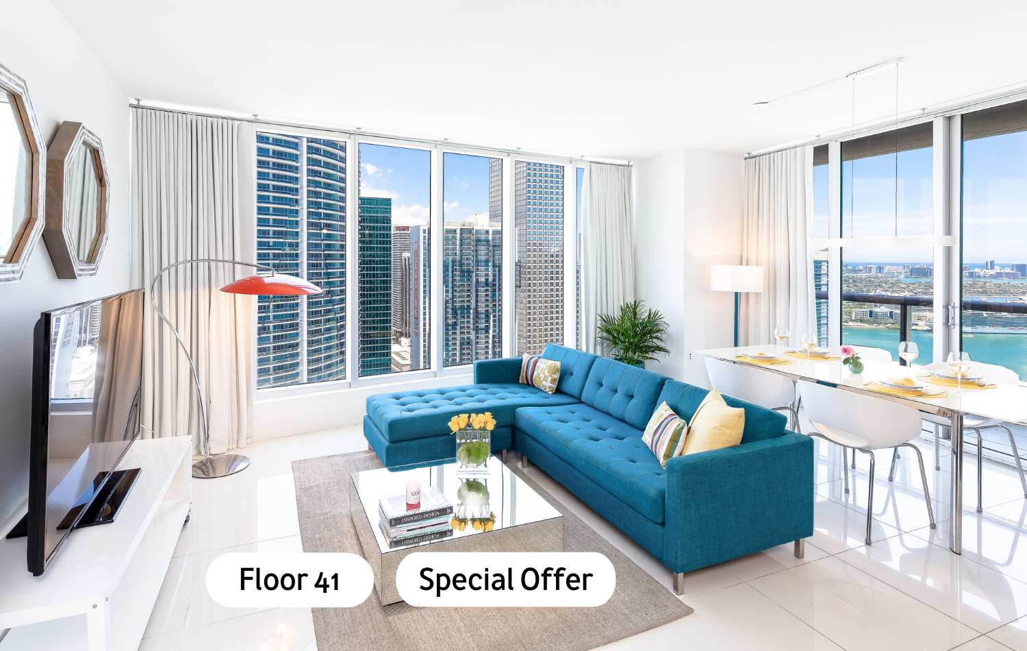 Bright living room featuring a large teal sectional sofa, a glass coffee table, and a white TV console, all set against a backdrop of expansive city skyscrapers and partial water views through floor-to-ceiling windows.