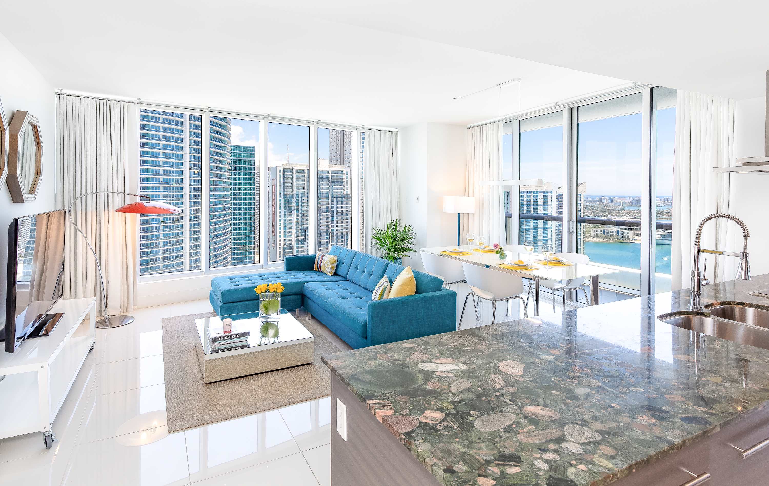 Open-concept living and dining area with a teal sectional sofa, glass coffee table, and a white dining table with chairs, all illuminated by natural light from large windows overlooking city and water views. A kitchen counter is visible in the foreground.