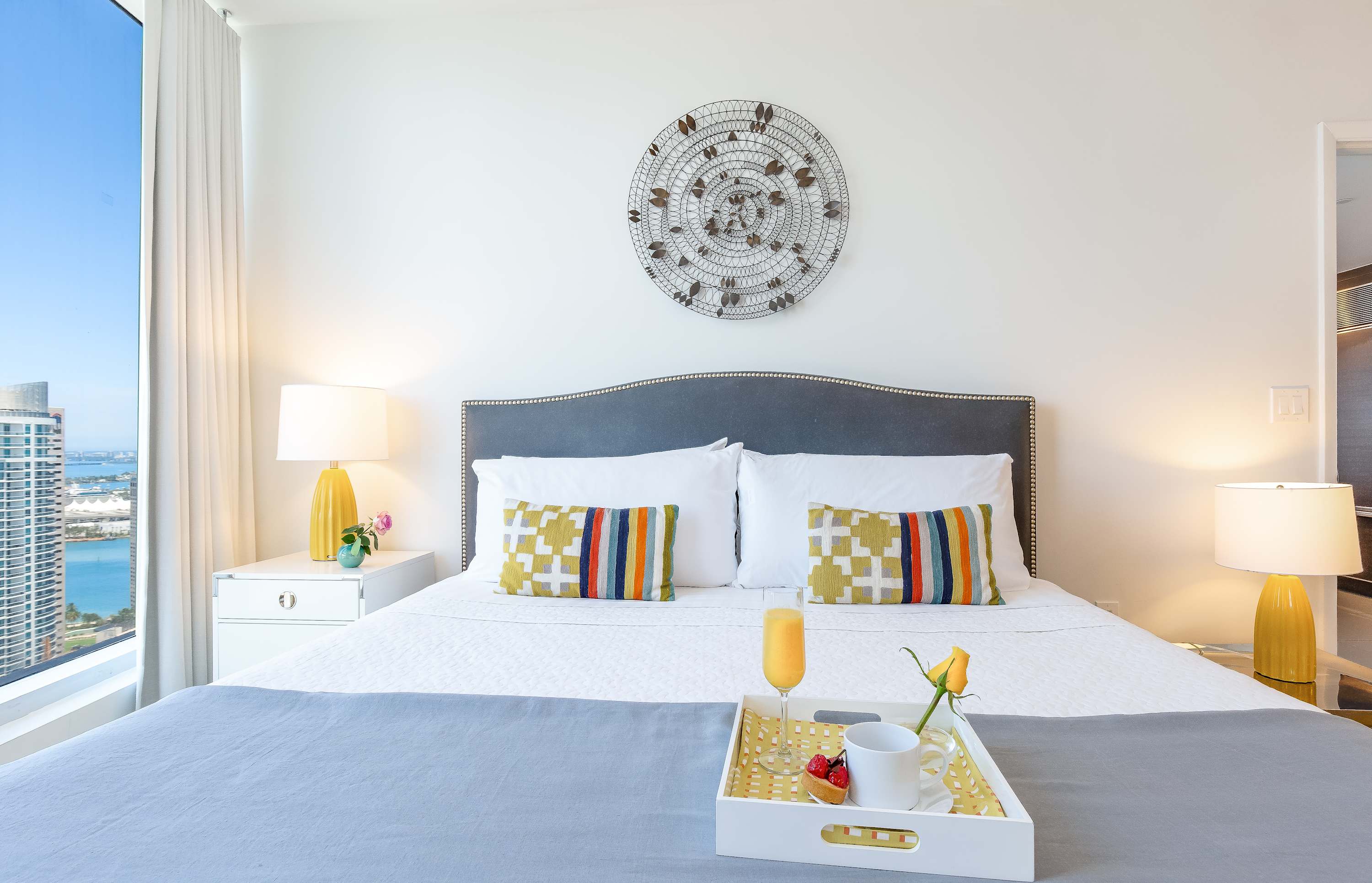Serene bedroom with a large bed, grey upholstered headboard, and vibrant throw pillows, flanked by white nightstands with yellow lamps. Large windows offer a glimpse of the city and water views.