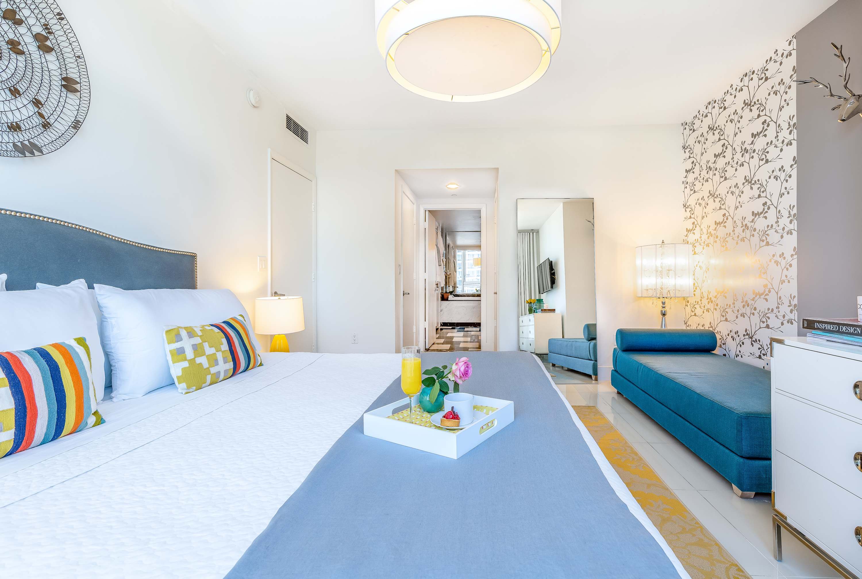 Bright bedroom with a large bed featuring a grey upholstered headboard, white bedding, and colorful throw pillows. A breakfast tray is placed on the bed, and a full-length mirror reflects the room.
