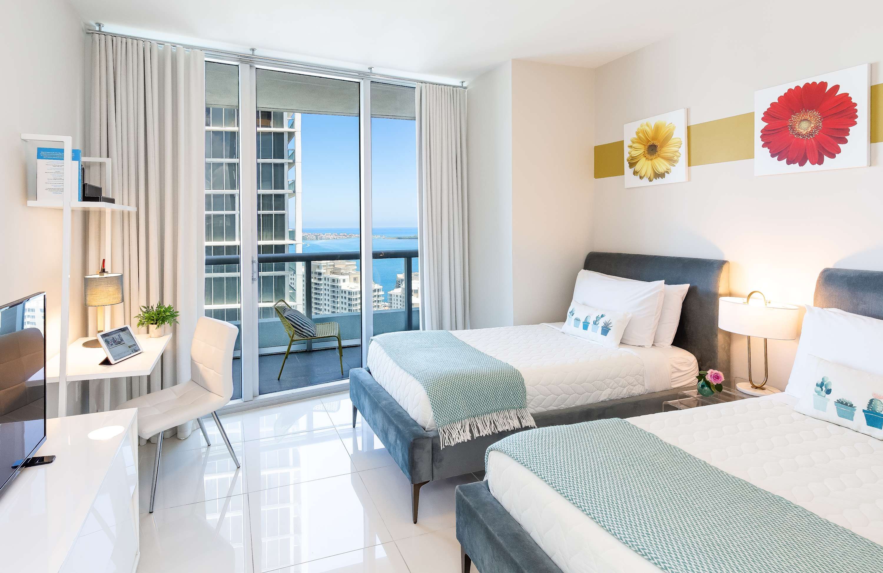 Bright bedroom with two twin beds featuring grey upholstered frames and light blue throws, a white desk with a chair, and a sliding glass door leading to a balcony with ocean views. Walls are white with yellow and red floral art.