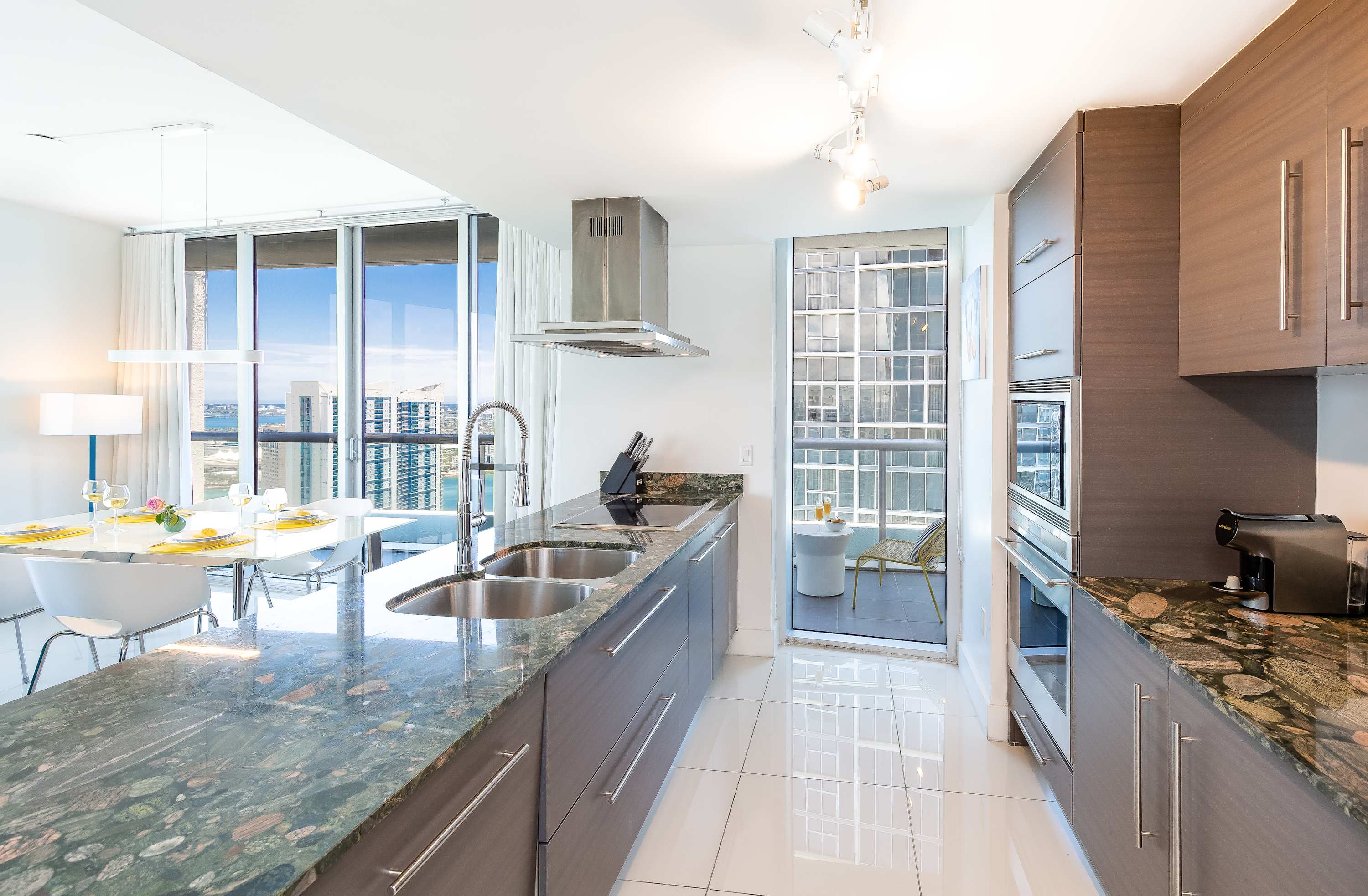 Modern kitchen with dark wood cabinetry, speckled green granite countertops, and stainless steel appliances, featuring a double sink and an open view towards the dining area and balcony with water views.