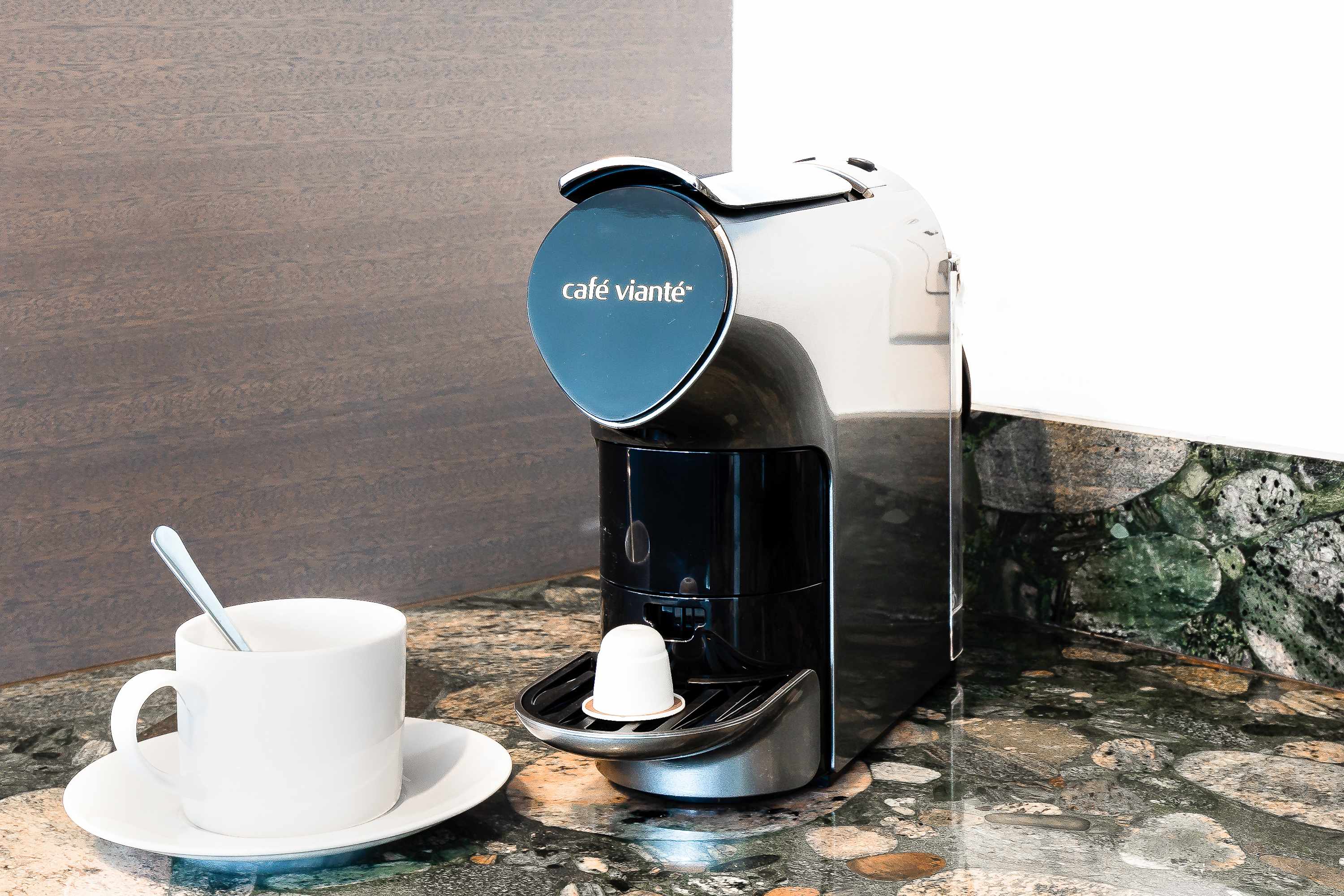 Close-up of a sleek, silver "Café Vianté" pod coffee machine on a speckled green granite countertop, with a white coffee cup and spoon ready for use.