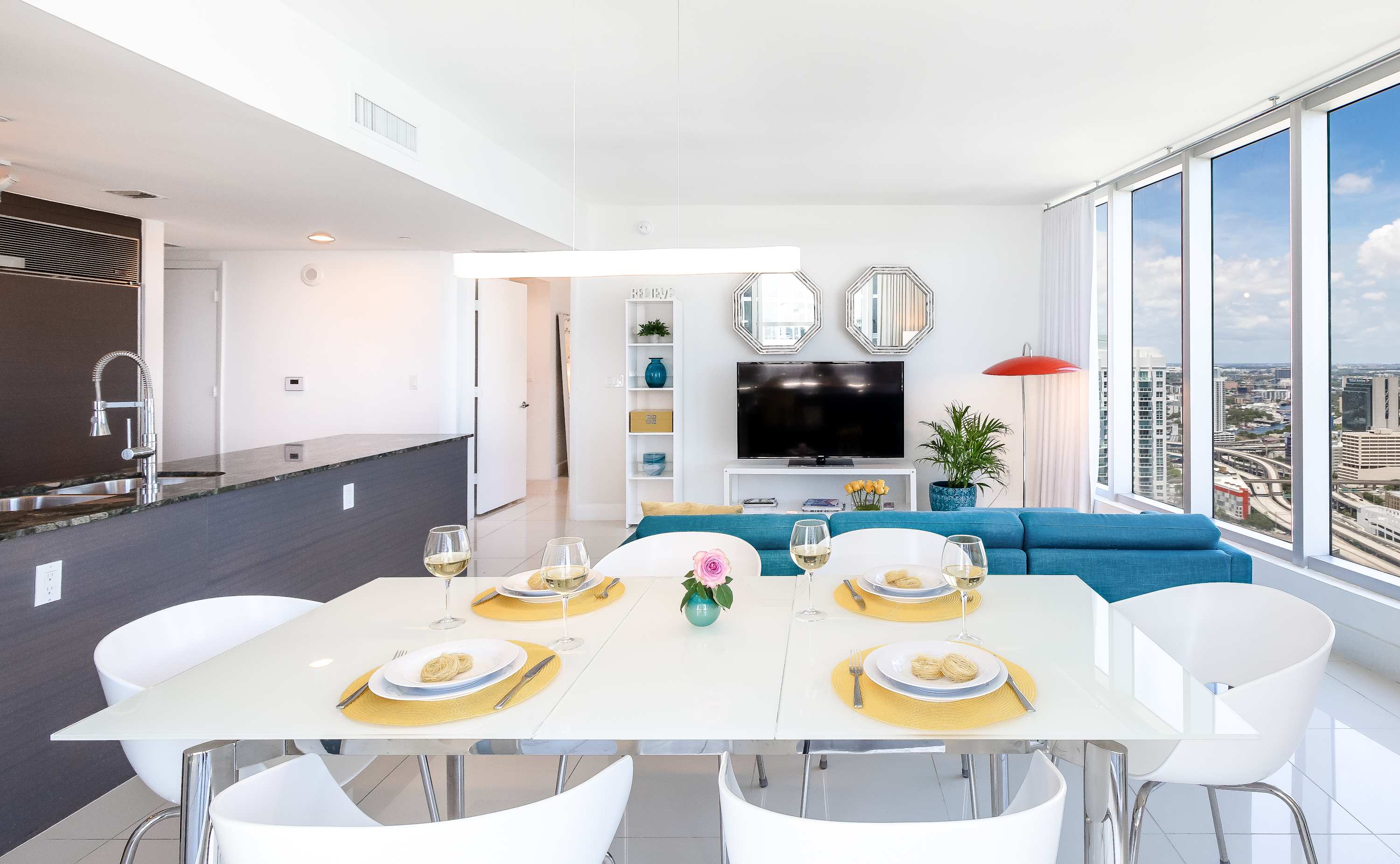 Elegant dining area featuring a white rectangular table set for four with yellow placemats and white chairs, positioned near large windows offering expansive city and water views. The living room with a blue sofa is visible in the background.