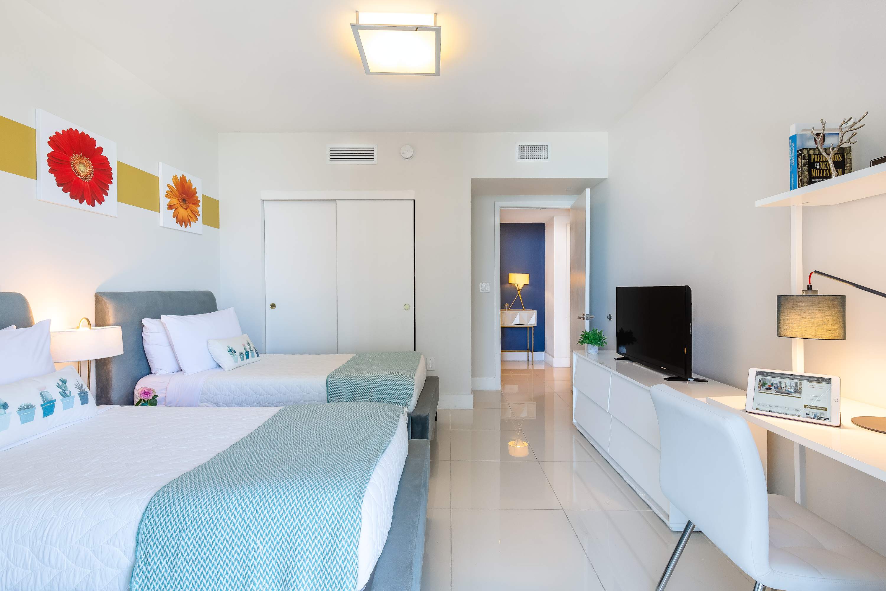 Modern bedroom with two twin beds, white tile flooring, and a white dresser with a flat-screen TV. A white desk and chair are visible on the right, and an open doorway leads to another room with blue walls.