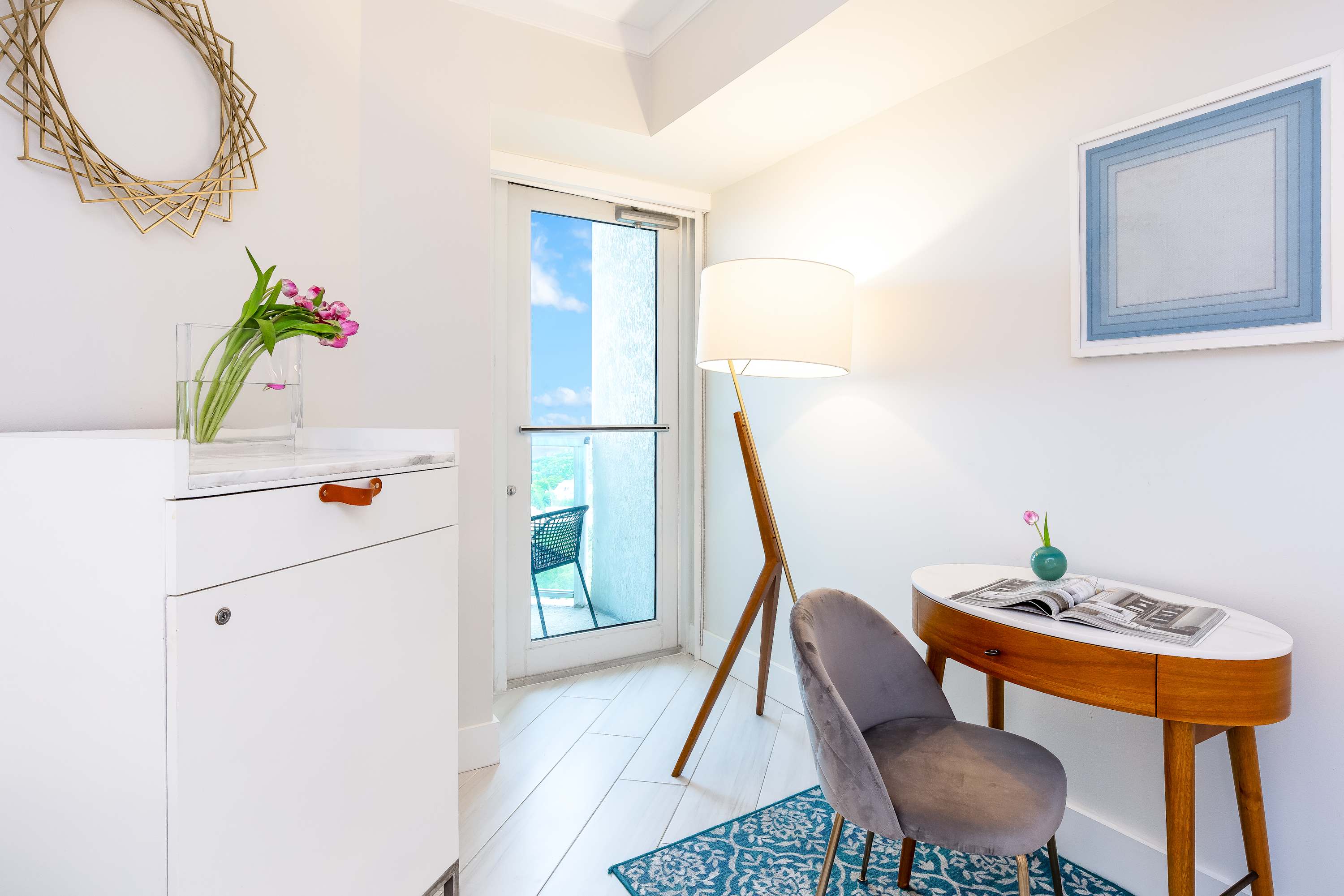 Cozy corner of the bedroom with a small wooden desk and a grey velvet chair, illuminated by a tripod floor lamp. A glass door provides access to a balcony, and a white cabinet stands nearby.