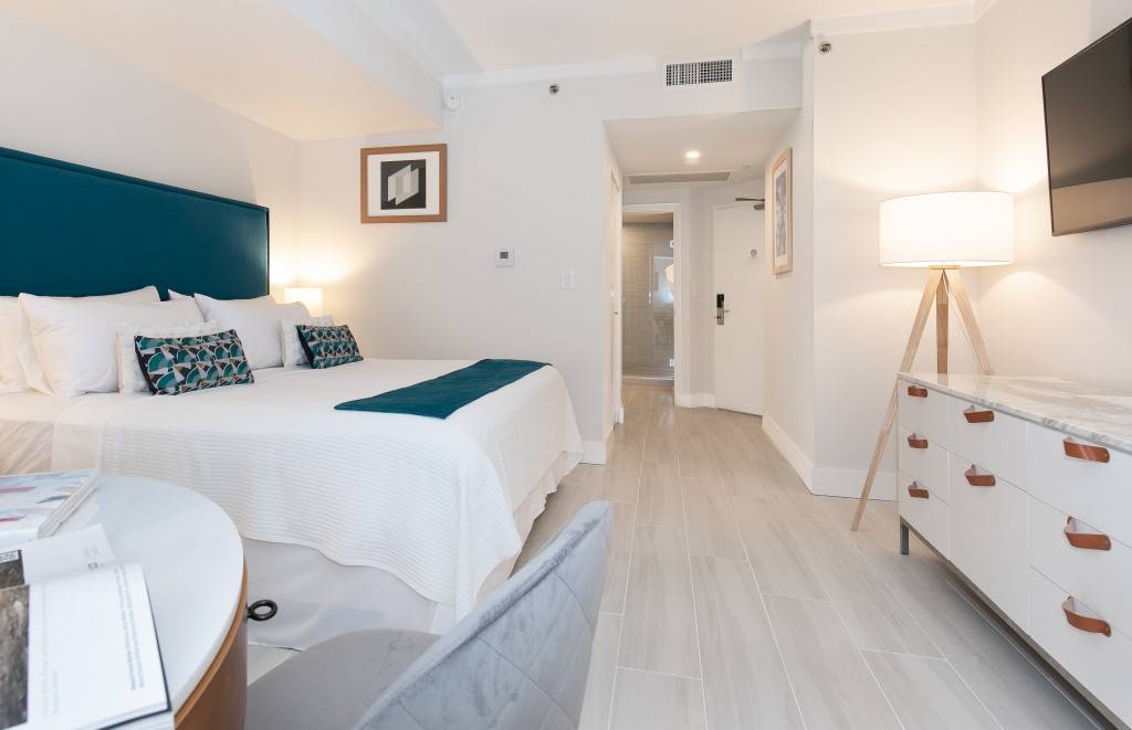 View of the bedroom from the foot of the bed, showing the teal upholstered headboard, white bedding, and light wood-look flooring. A doorway leads to the bathroom, and a dresser with a TV is visible on the right.
