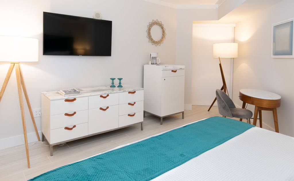 Modern bedroom area featuring a white dresser with a marble top and brown leather pulls, topped with a flat-screen TV. A tripod floor lamp and a decorative gold mirror adorn the white wall.