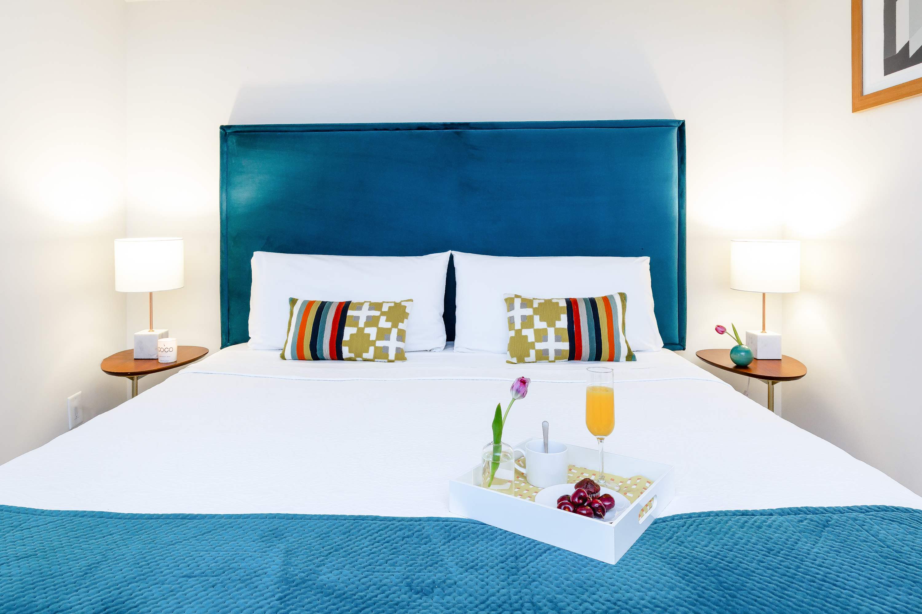 Close-up of the king bed with a teal upholstered headboard, crisp white bedding, and colorful geometric throw pillows. Two modern nightstands with lamps flank the bed, which holds a breakfast tray.