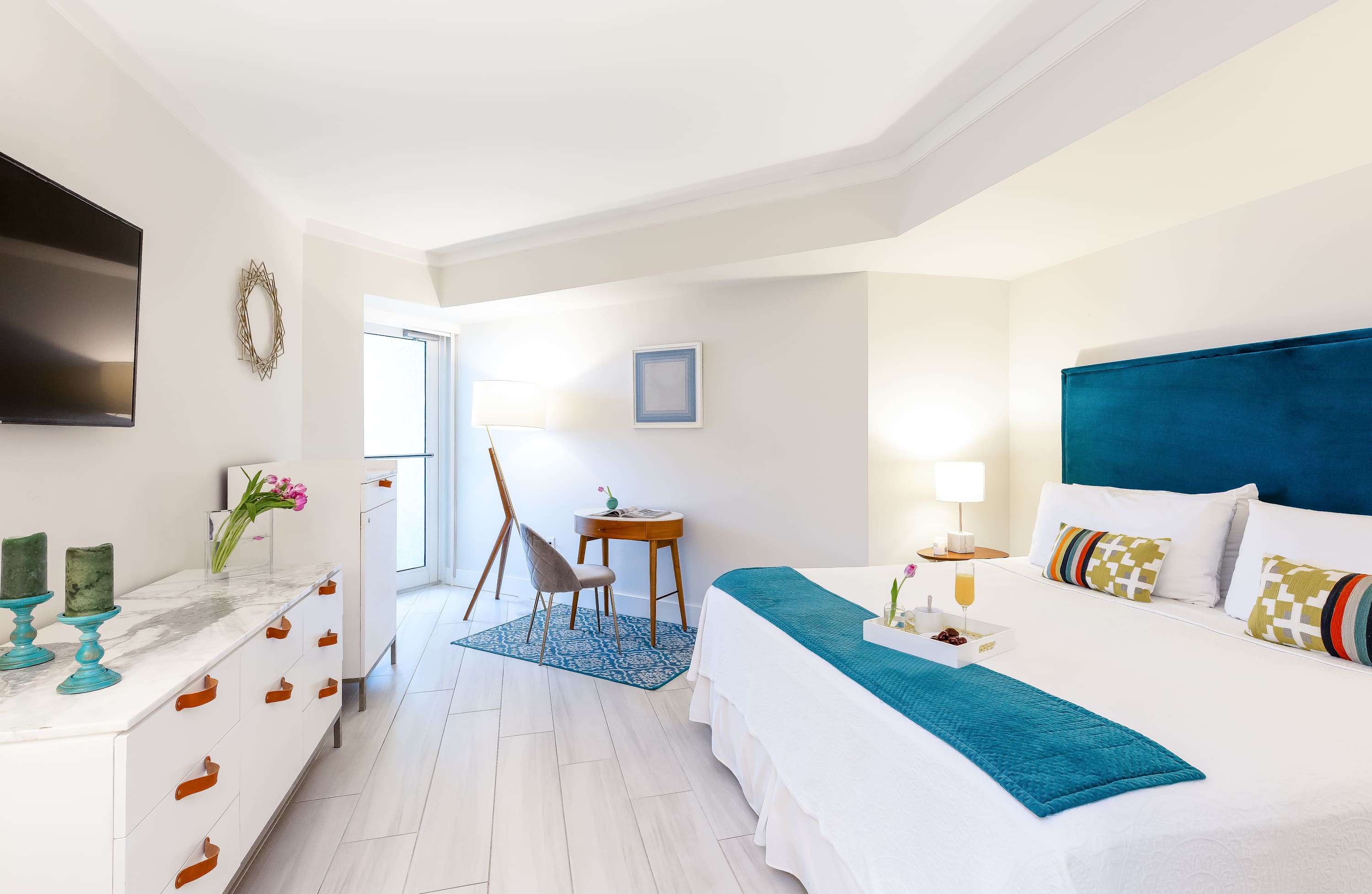 Spacious bedroom with light wood-look flooring and white walls, featuring a king bed with a teal headboard, a white dresser with a marble top and TV, and a small desk area near a balcony door.