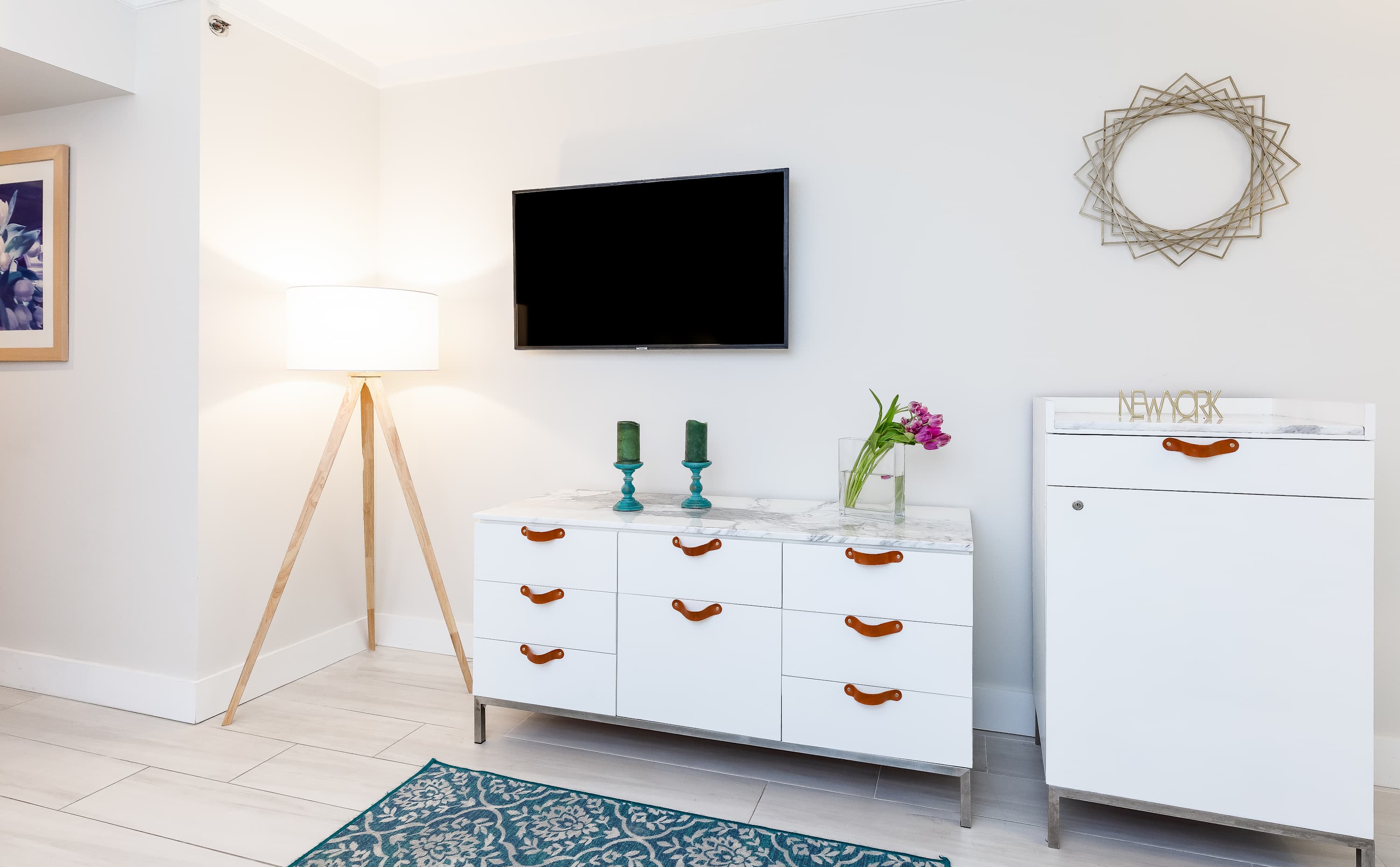 Modern bedroom space featuring a white dresser with a marble top and brown leather pulls, positioned beneath a wall-mounted flat-screen TV. A tripod floor lamp and a decorative gold mirror add to the contemporary aesthetic.