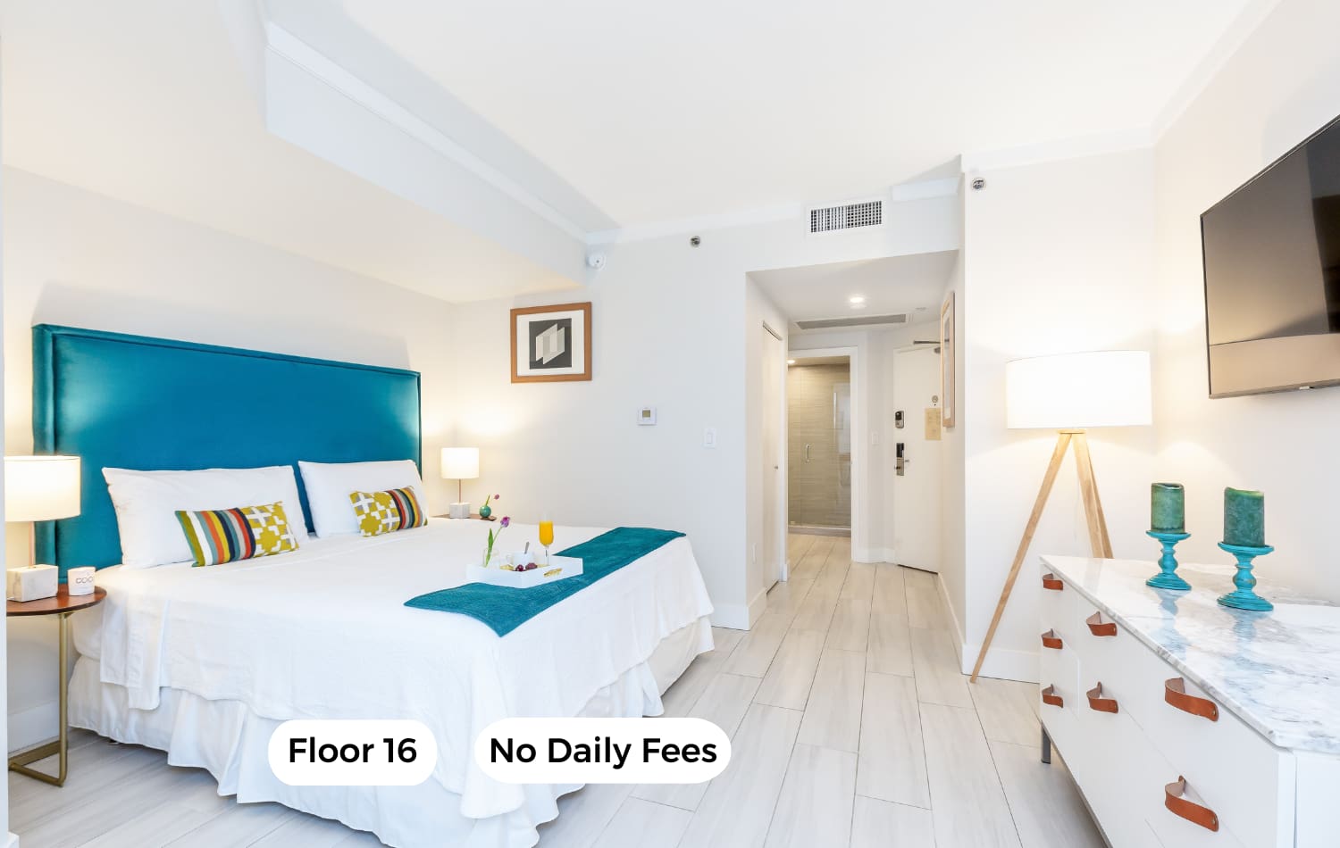 Bright bedroom featuring a king bed with a teal upholstered headboard, white bedding, and a teal throw. Light wood-look flooring, white walls, and a white dresser with a marble top and a wall-mounted TV complete the modern look.