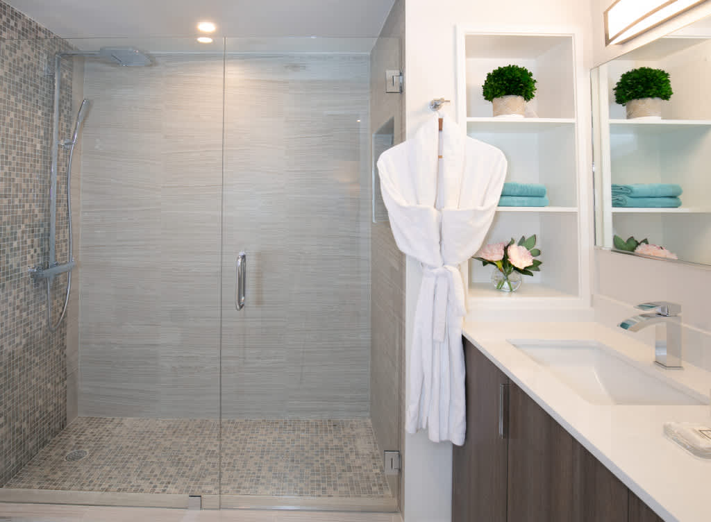 Contemporary bathroom with a spacious walk-in shower featuring a glass door, mosaic tile floor, and light grey wall tiles. A vanity with a white countertop, sink, and open shelving for towels and decor completes the space.