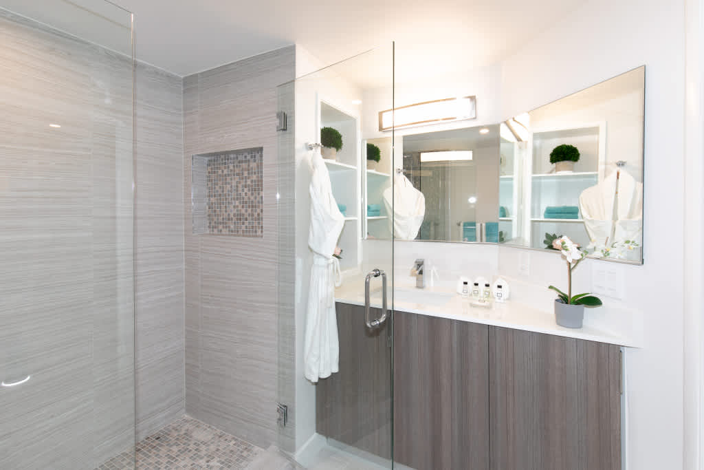 Modern bathroom featuring a glass-enclosed shower with light wood-grain tiled walls and a mosaic-tiled niche, alongside a vanity with a white countertop, wood-look cabinetry, and a large mirror.