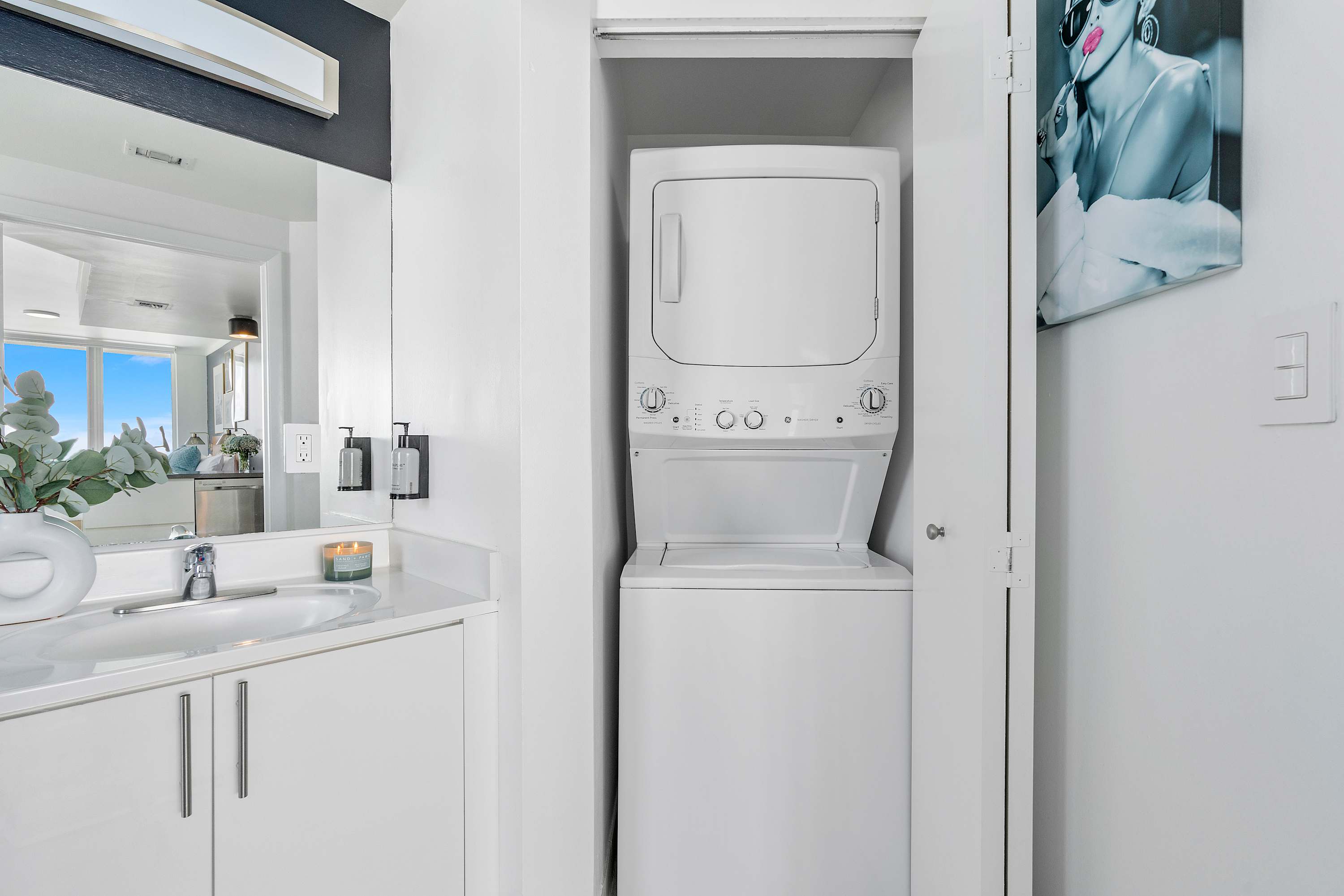 Clean bathroom vanity with a white countertop, single sink, and a large mirror, adjacent to a closet housing a stacked white washer and dryer unit. A decorative plant and candle are on the counter.