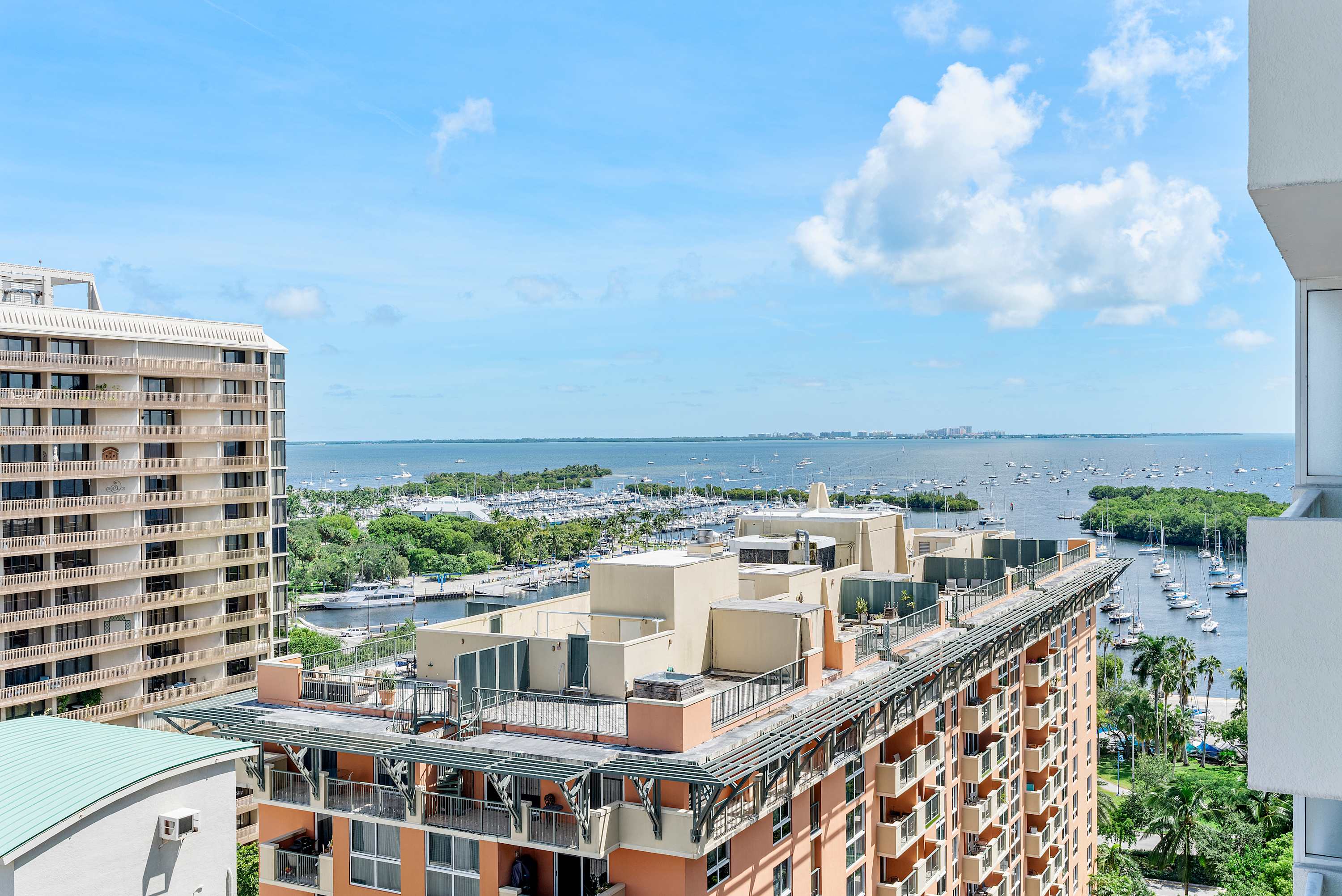 A panoramic high-rise view of a vibrant bay dotted with numerous boats and yachts, framed by city buildings and lush green areas under a bright blue sky with scattered clouds.