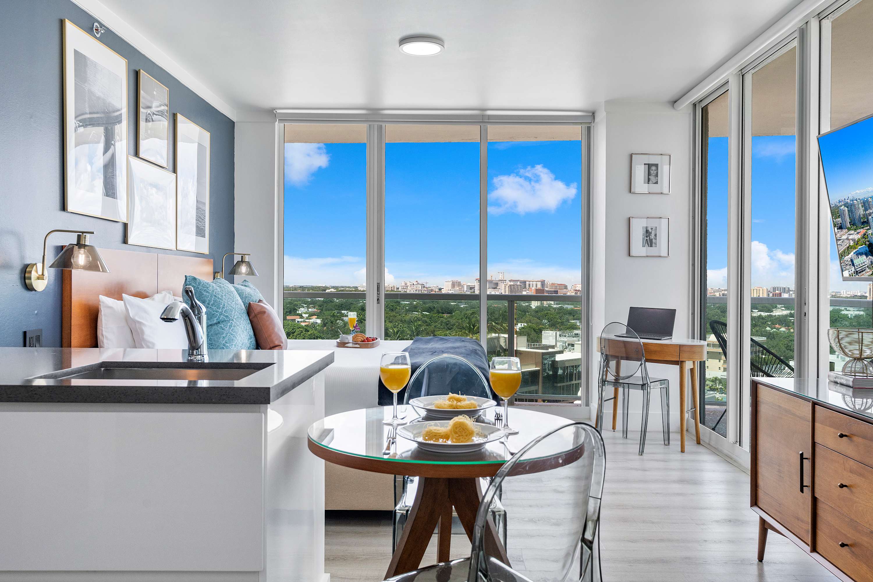 A bright studio apartment featuring a king bed, a small dining table with clear chairs, and a compact kitchen area. Large windows offer a panoramic view of the city and distant greenery, enhancing the open feel.
