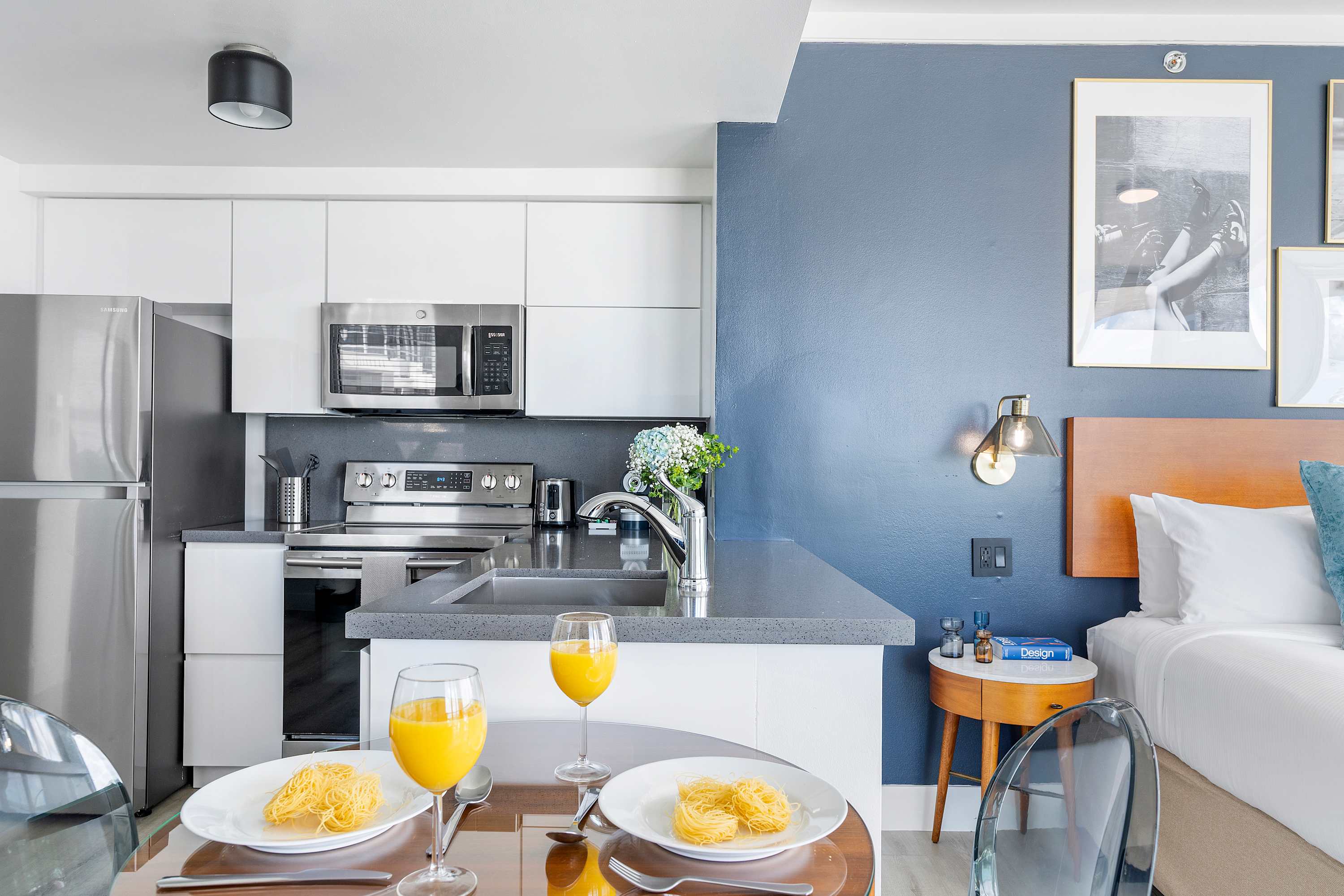 A modern kitchen area featuring sleek white flat-panel cabinets, stainless steel appliances, and a dark grey countertop. A small glass-top dining table with clear chairs is positioned in the foreground, ready for a meal.
