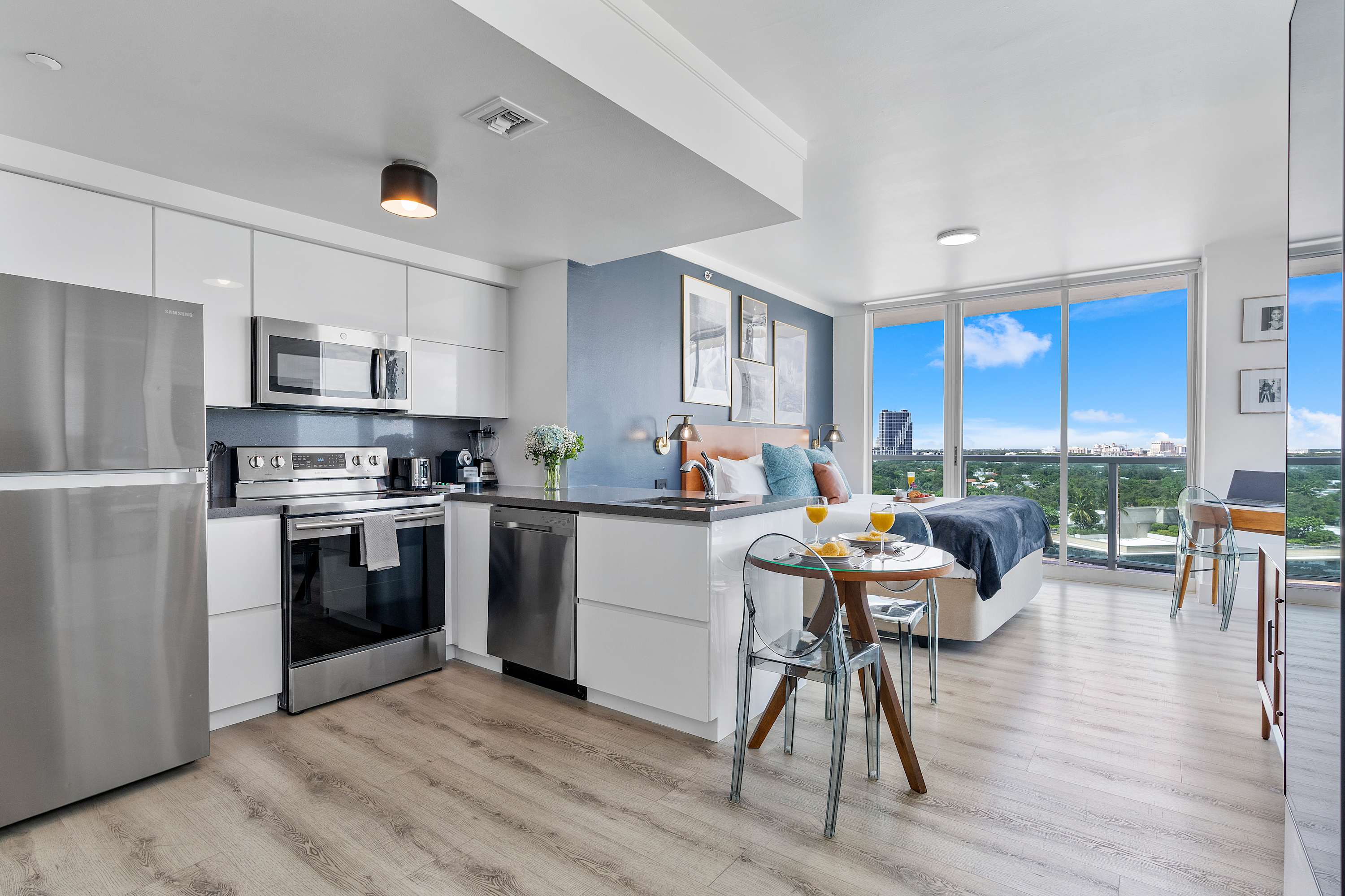 An open-concept studio apartment showcasing a modern kitchen, a small dining area with a glass table, and a comfortable king-sized bed. Floor-to-ceiling windows provide expansive city views, filling the space with natural light.