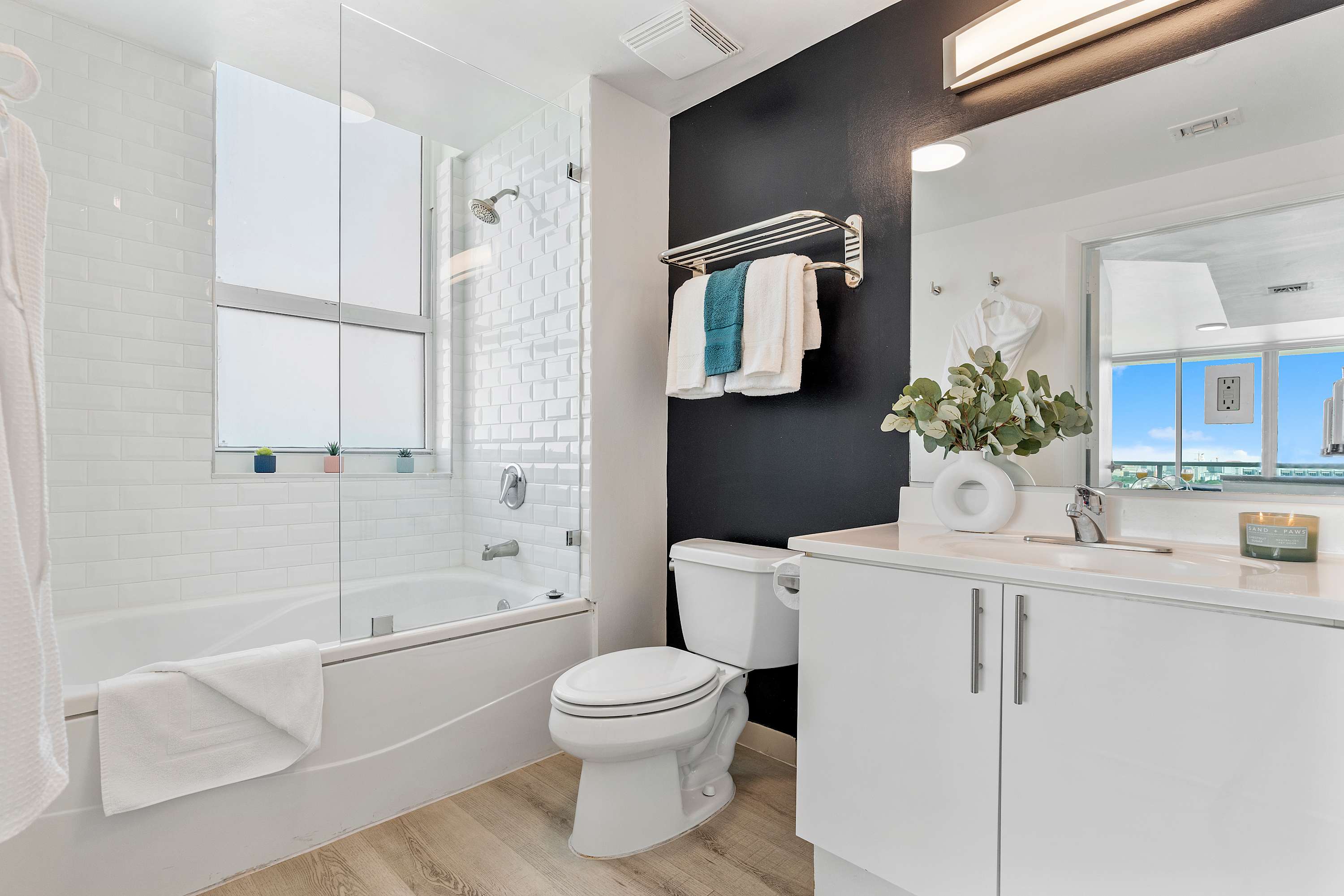Contemporary bathroom with a white bathtub and shower combo featuring subway tiles, a white toilet, and a vanity with a single sink. A dark accent wall behind the toilet provides contrast.