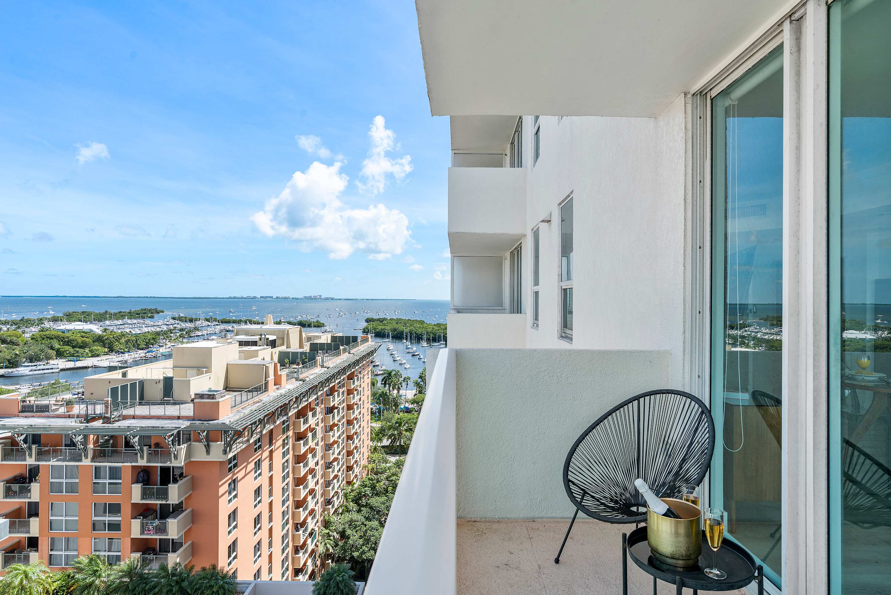 A private balcony offering a stunning sea view of a bay filled with numerous boats and yachts, framed by surrounding city buildings. A stylish black wire chair and a small table with champagne are set up for relaxation.