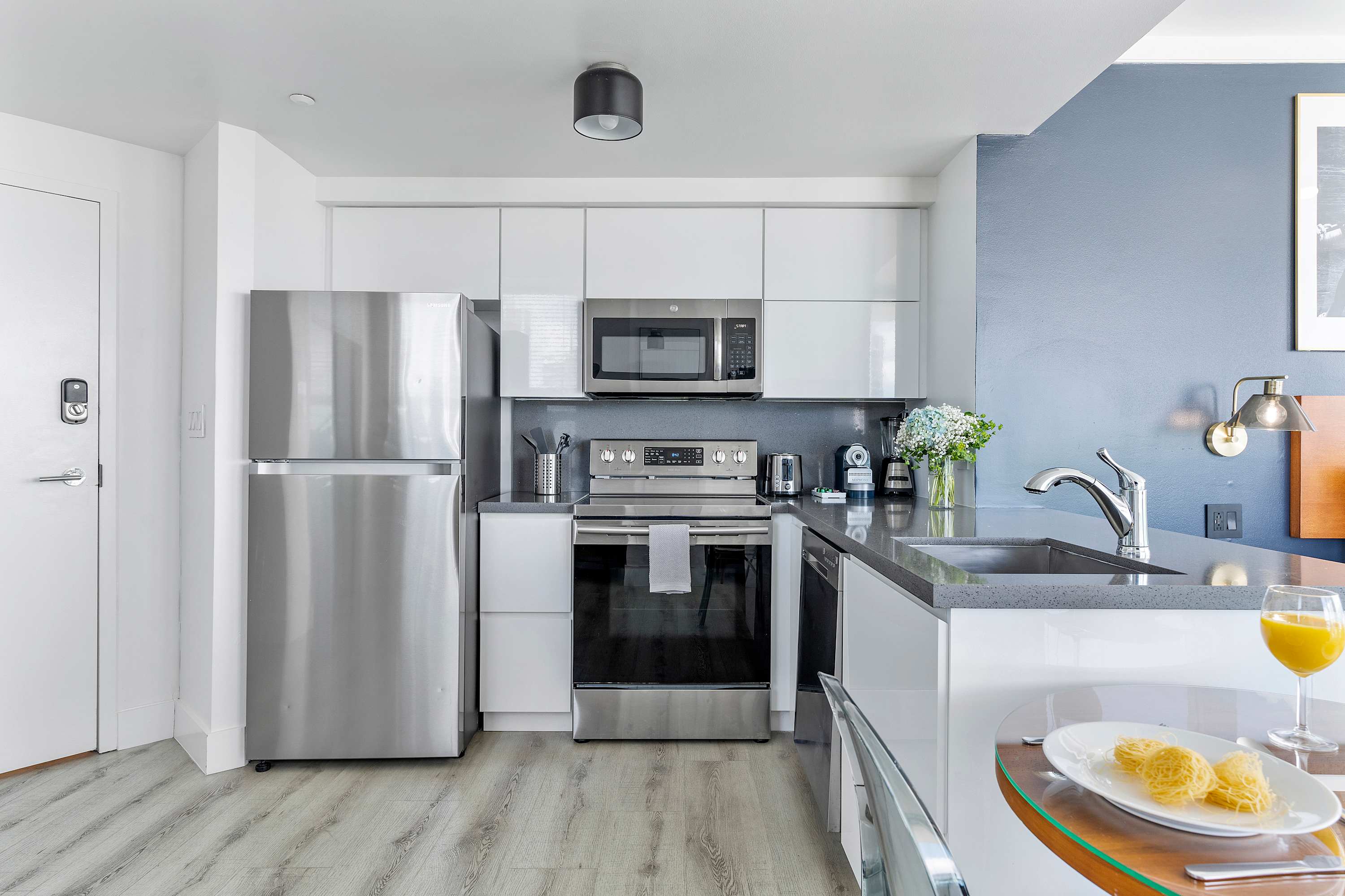 A contemporary kitchen equipped with a full suite of stainless steel appliances, including a refrigerator, oven, microwave, and dishwasher. The space features white cabinetry, a dark grey countertop, and light hardwood flooring.