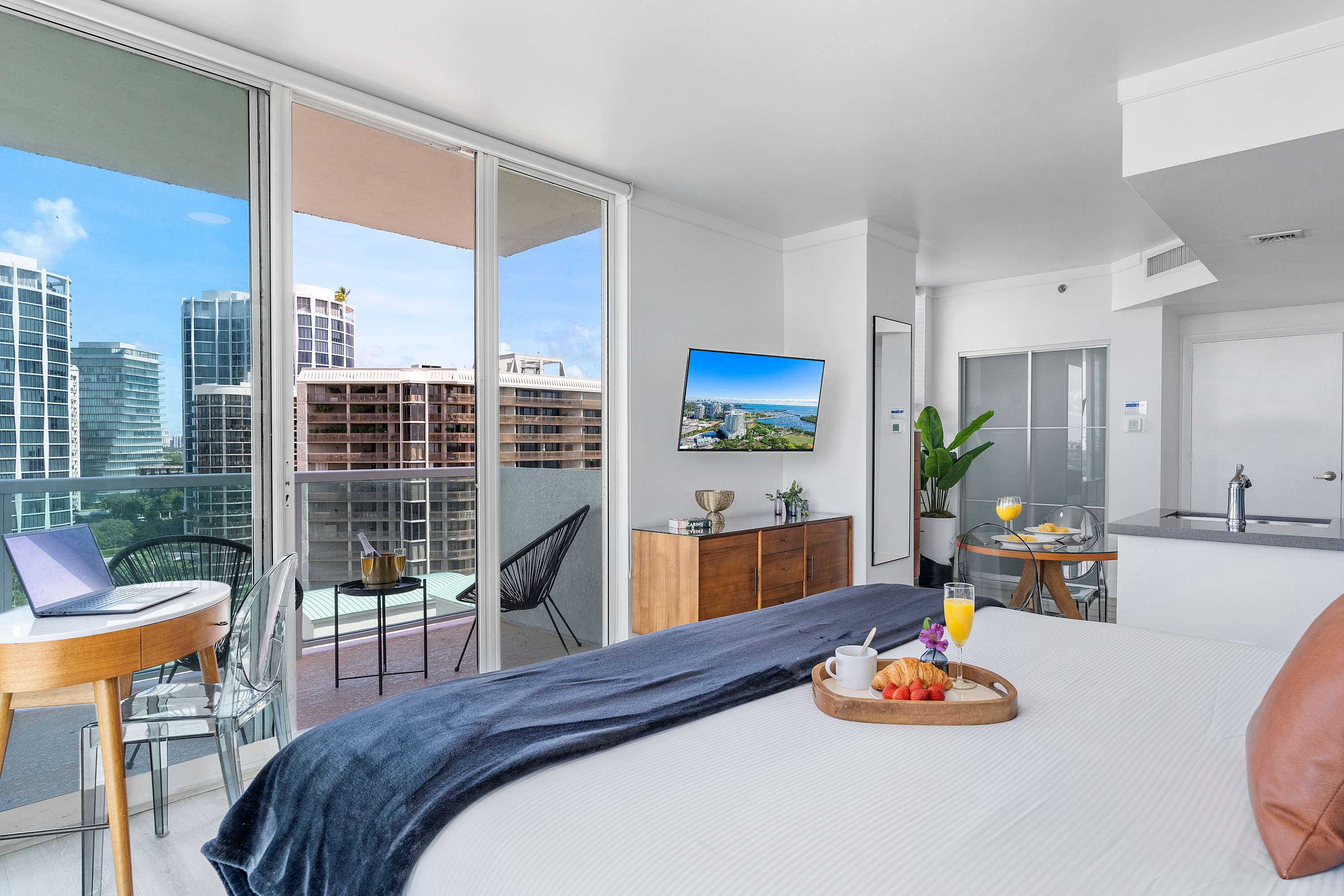 The studio's sleeping area features a king bed with a dark blue throw, a small wooden desk with a laptop, and direct access to a private balcony through sliding glass doors. A flat-screen TV is mounted on the wall.