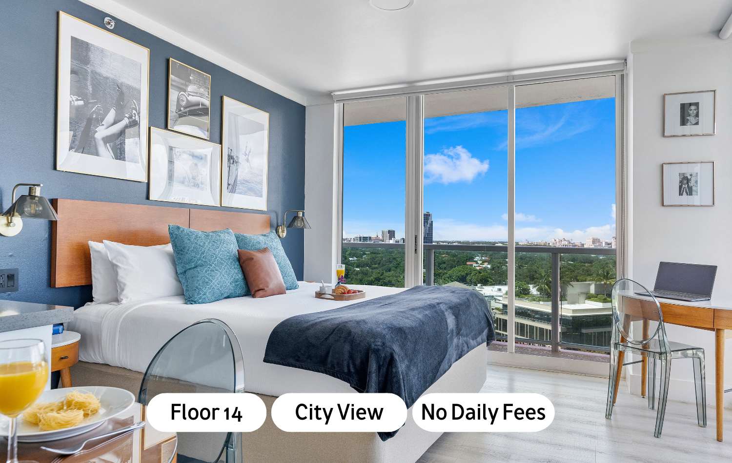 A modern studio bedroom featuring a king-sized bed with a blue accent wall and floor-to-ceiling windows offering expansive city views. A small desk and chair are positioned by the window, with a breakfast tray on the bed.