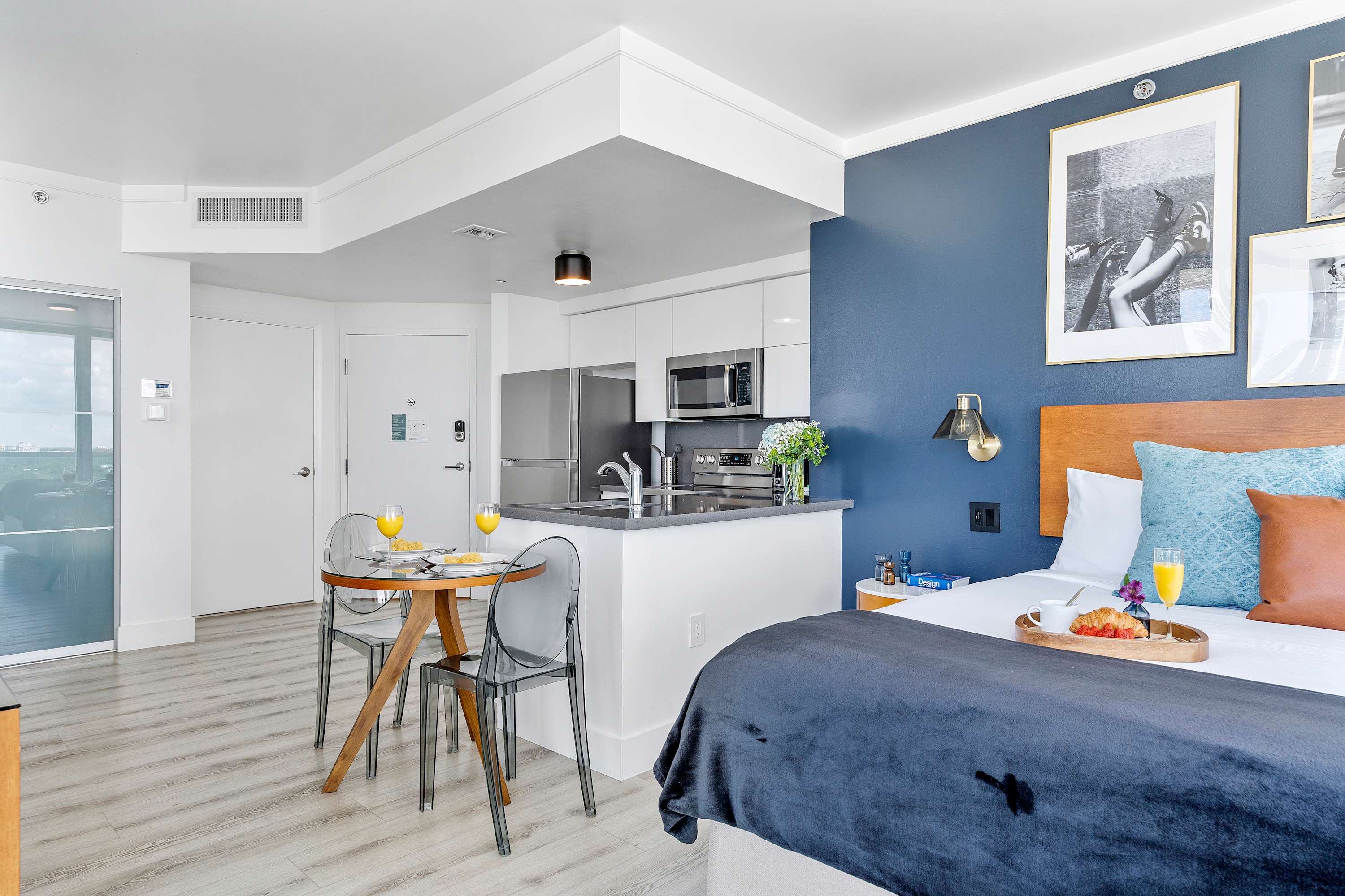 Bright studio apartment featuring a queen bed with a blue duvet, a compact kitchen with white cabinetry and stainless steel appliances, and a small dining area with a round glass table and clear chairs. A dark blue accent wall adds a pop of color.