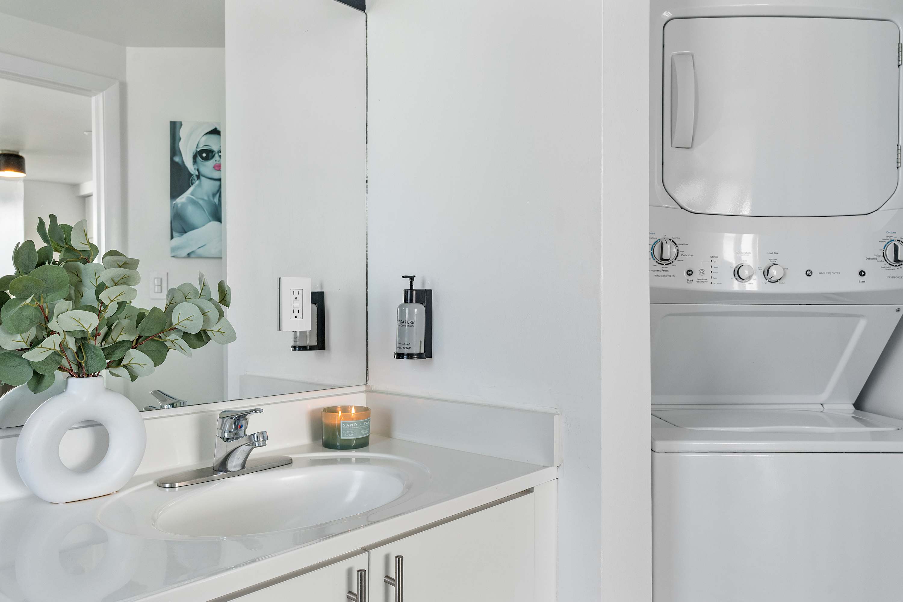 Modern bathroom vanity with a white countertop, single sink, large mirror, and a stacked white washer and dryer unit in the background. A white vase with eucalyptus branches adds a touch of greenery.